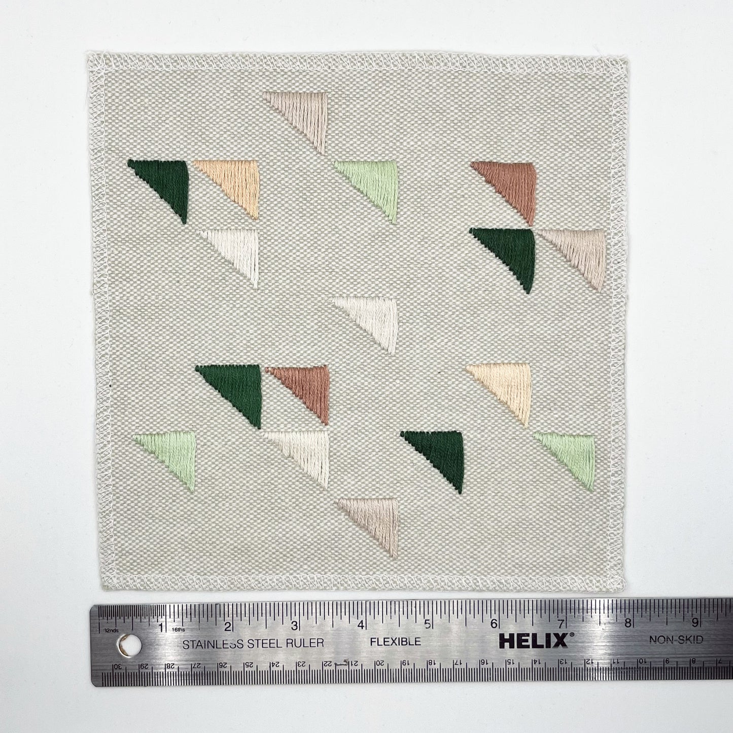 Large square natural colored patch embroidered with randomly placed triangles in peaches, mauves and greens, next to a metal ruler showing a width of 8 inches, on a white background
