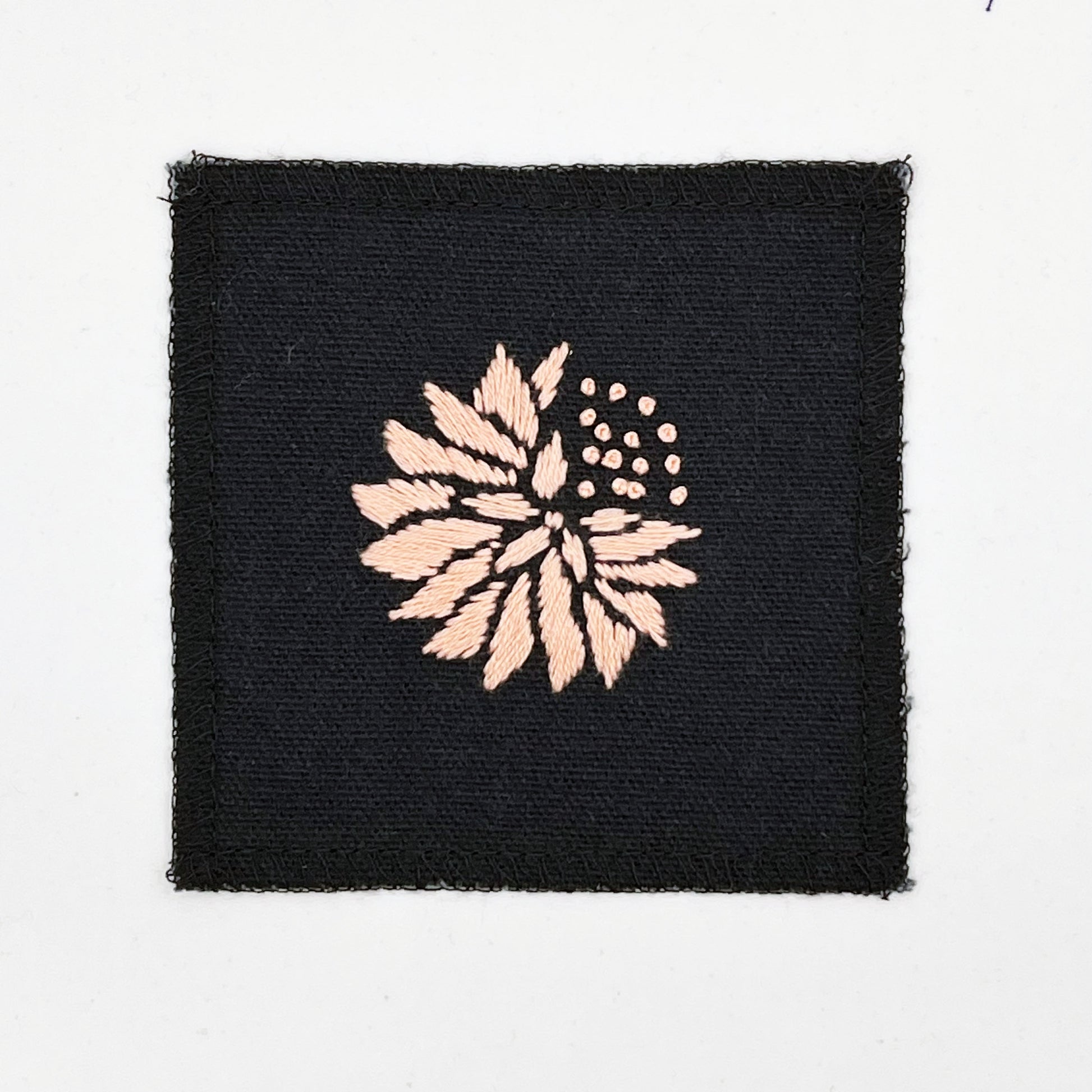 a square patch in black fabric, hand embroidered with a peach colored abstract dandelion made mostly of petals, with a quarter of them replaced with french knots, with overlocked edges, on a white background