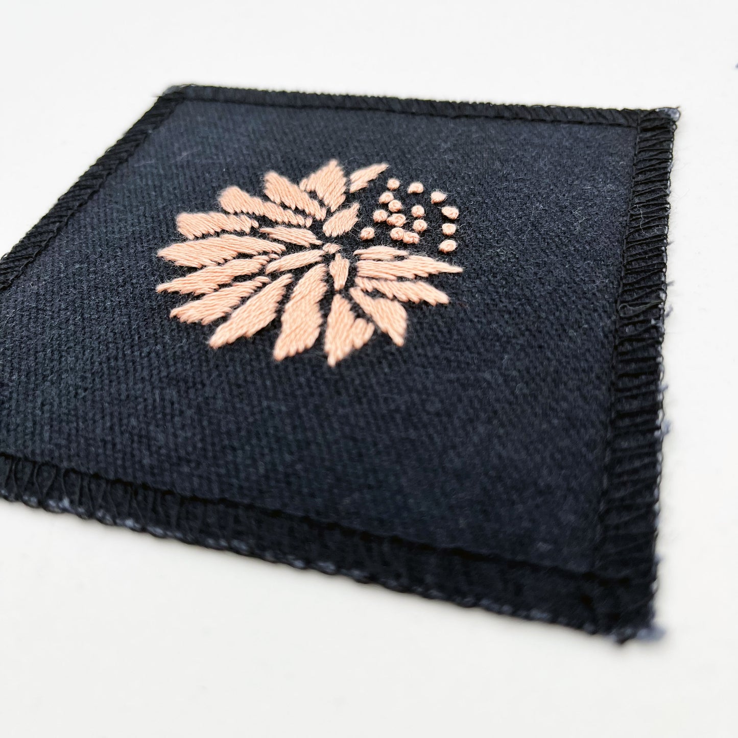 a close up angled view of a square patch in black fabric, hand embroidered with a peach colored abstract dandelion made mostly of petals, with a quarter of them replaced with french knots, with overlocked edges, on a white background