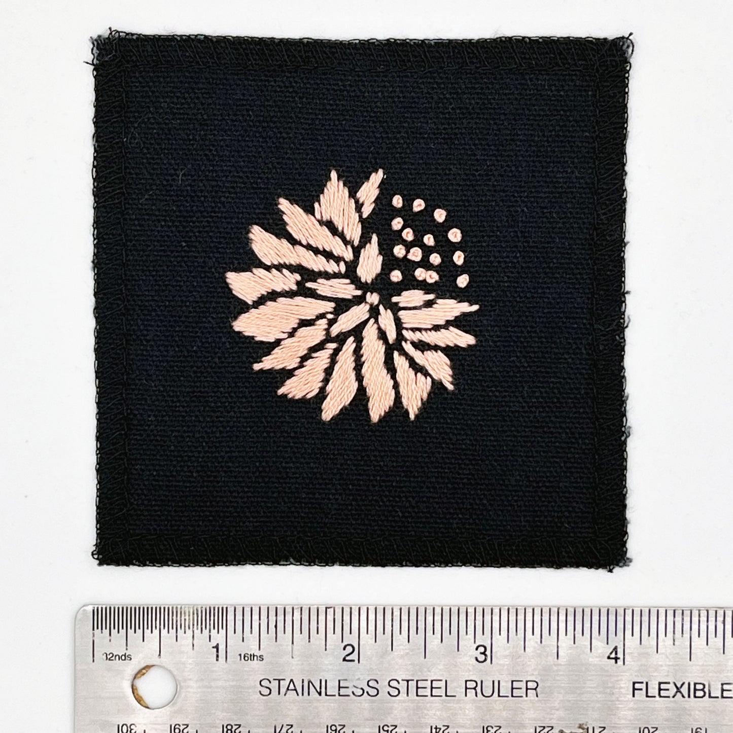 a square patch in black fabric, hand embroidered with a peach colored abstract dandelion made mostly of petals, with a quarter of them replaced with french knots, with overlocked edges, next to a metal ruler to show a width of four inches