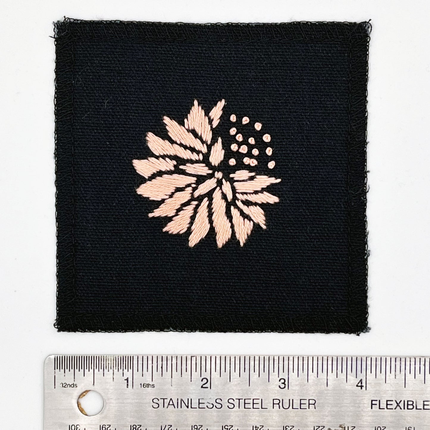 a square patch in black fabric, hand embroidered with a peach colored abstract dandelion made mostly of petals, with a quarter of them replaced with french knots, with overlocked edges, next to a metal ruler to show a width of four inches