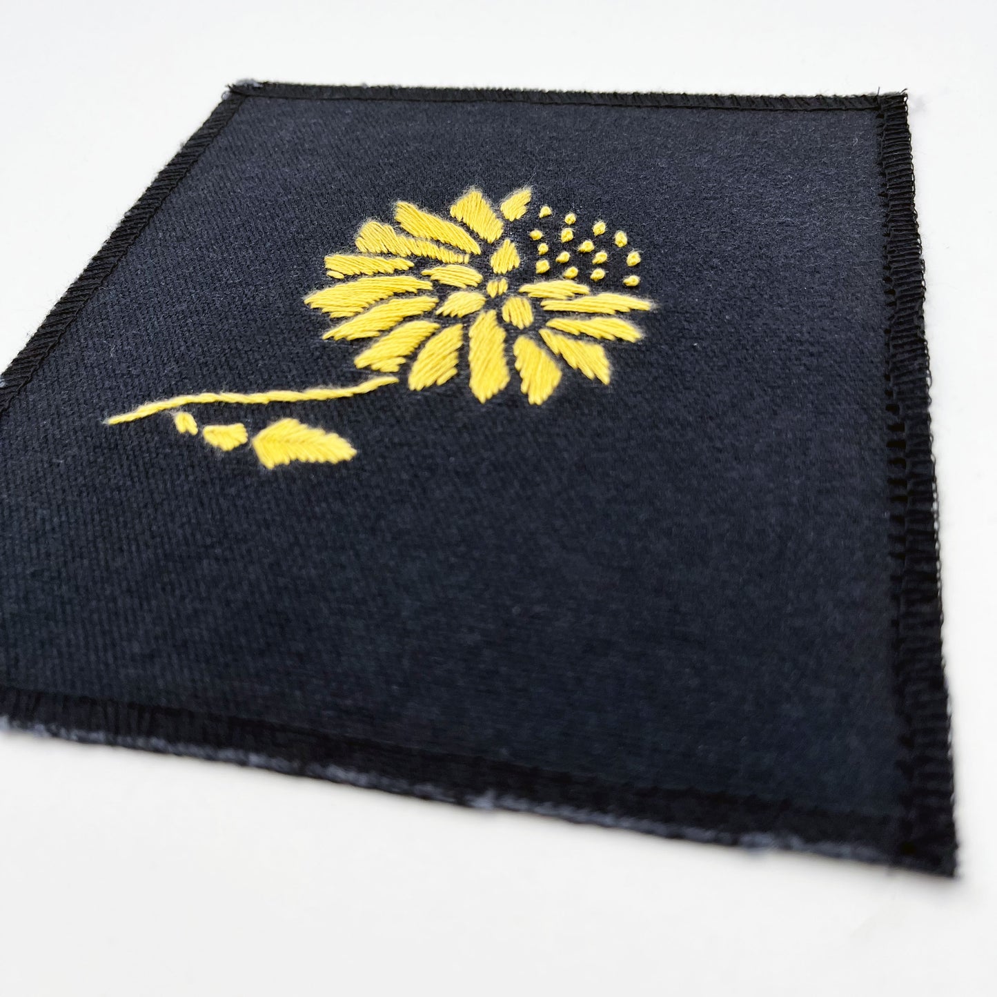 a close up angled view of a square patch in black fabric, hand embroidered with a yellow gold colored abstract dandelion made mostly of petals, with a quarter of them replaced with french knots, with overlocked edges, on a white background