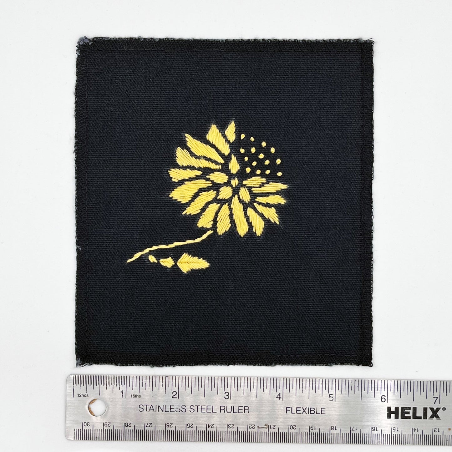 a square patch in black fabric, hand embroidered with a yellow gold colored abstract dandelion made mostly of petals, with a quarter of them replaced with french knots, with overlocked edges, next to a metal ruler to show a width of six inches