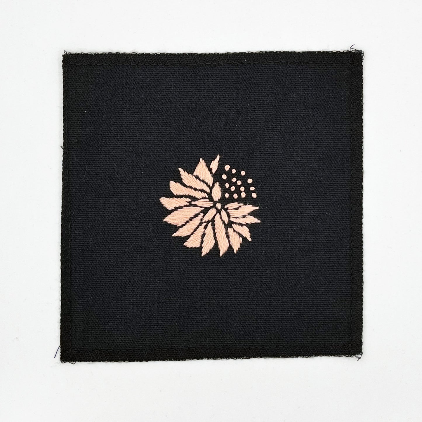 a square patch in black fabric, hand embroidered with a peach colored abstract dandelion made mostly of petals, with a quarter of them replaced with french knots, with overlocked edges, on a white background