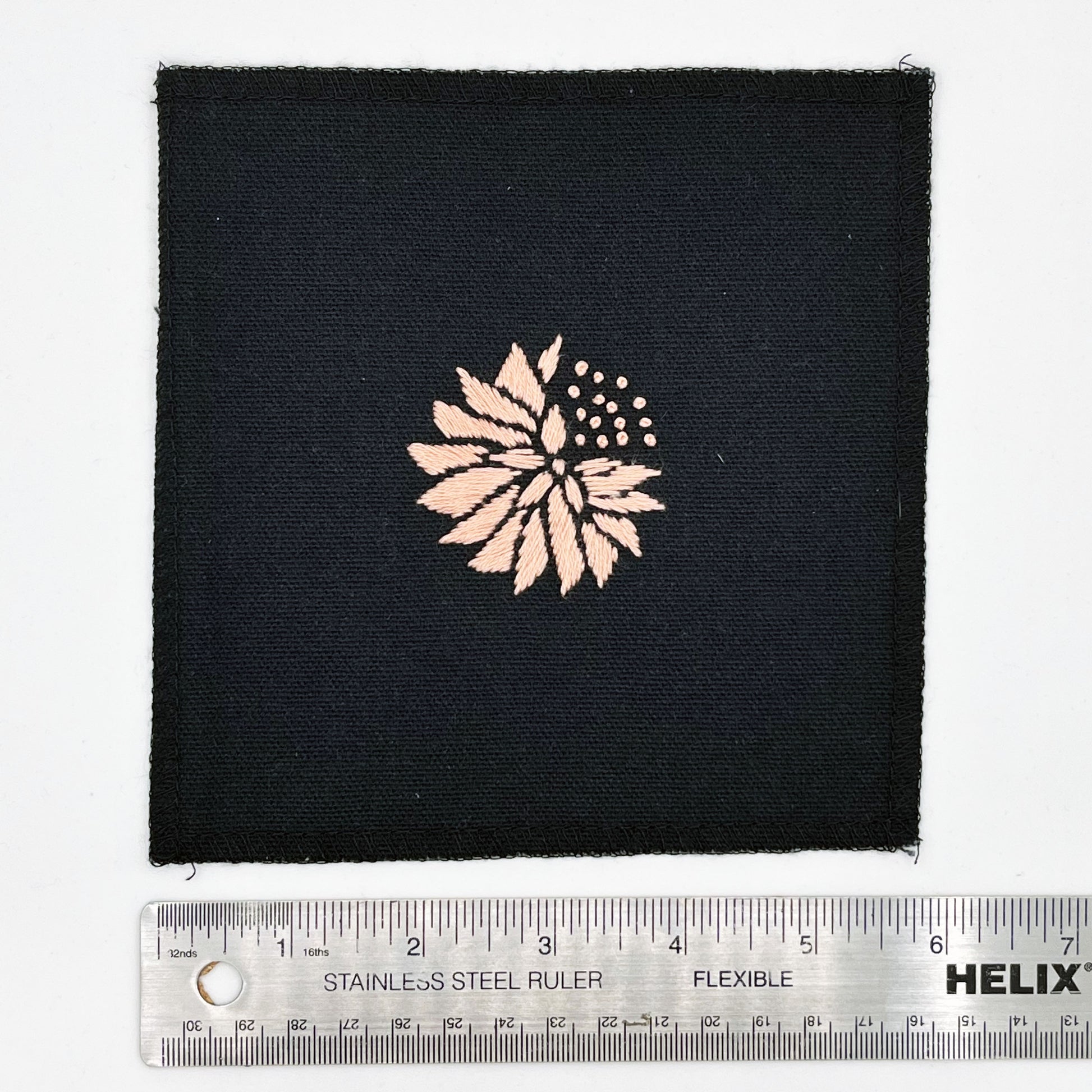 a square patch in black fabric, hand embroidered with a peach colored abstract dandelion made mostly of petals, with a quarter of them replaced with french knots, with overlocked edges, next to a metal ruler to show a width of six inches