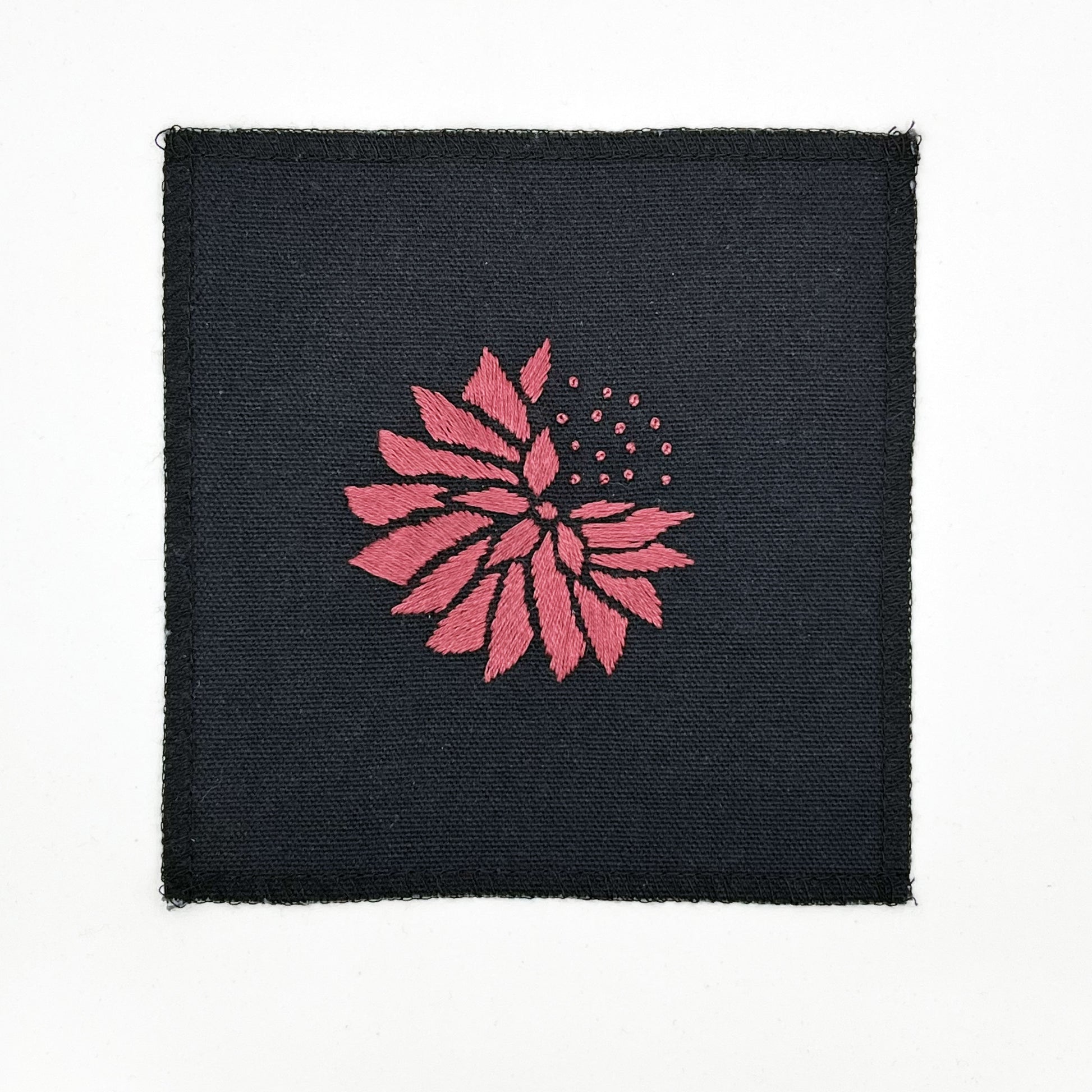 a square patch in black fabric, hand embroidered with a plum colored abstract dandelion made mostly of petals, with a quarter of them replaced with french knots, with overlocked edges, on a white background
