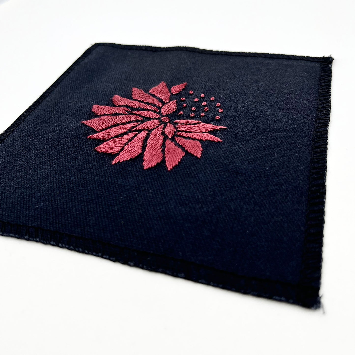 a close up angled view of a square patch in black fabric, hand embroidered with a plum colored abstract dandelion made mostly of petals, with a quarter of them replaced with french knots, with overlocked edges, on a white background