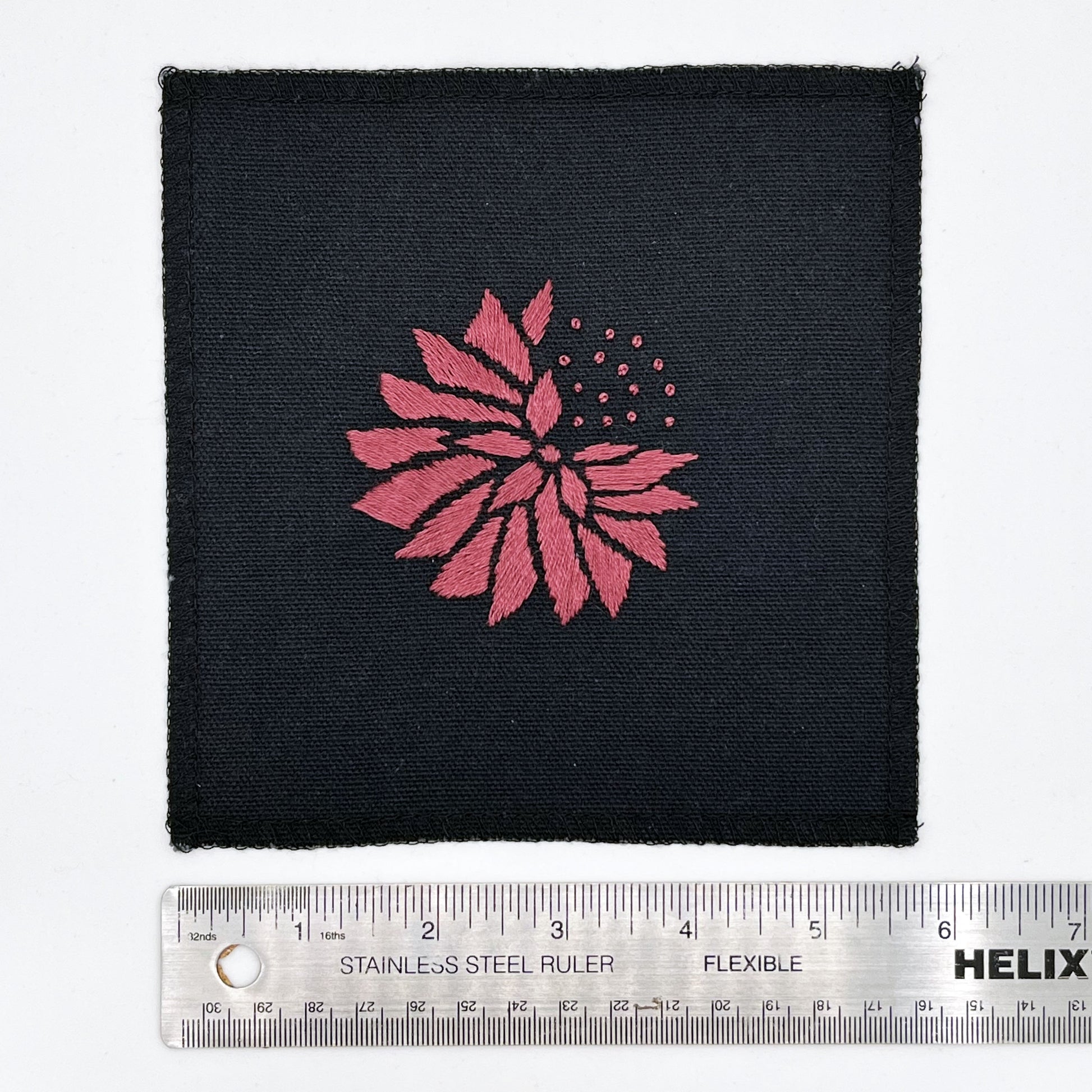 a square patch in black fabric, hand embroidered with a plum colored abstract dandelion made mostly of petals, with a quarter of them replaced with french knots, with overlocked edges, next to a metal ruler to show a width of six inches