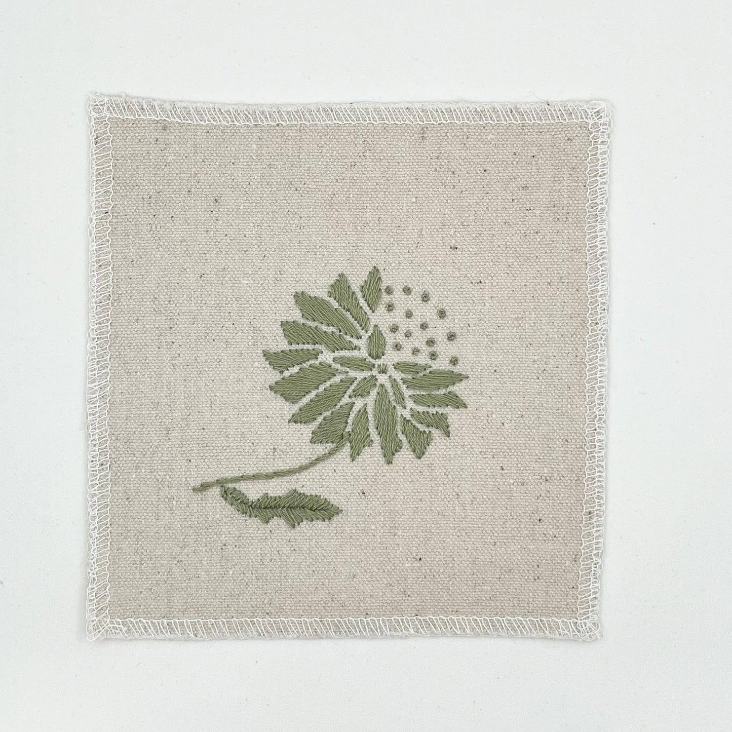 a square patch in natural colored fabric, hand embroidered with a sage colored abstract dandelion made mostly of petals, with a quarter of them replaced with french knots, with overlocked edges, on a white background