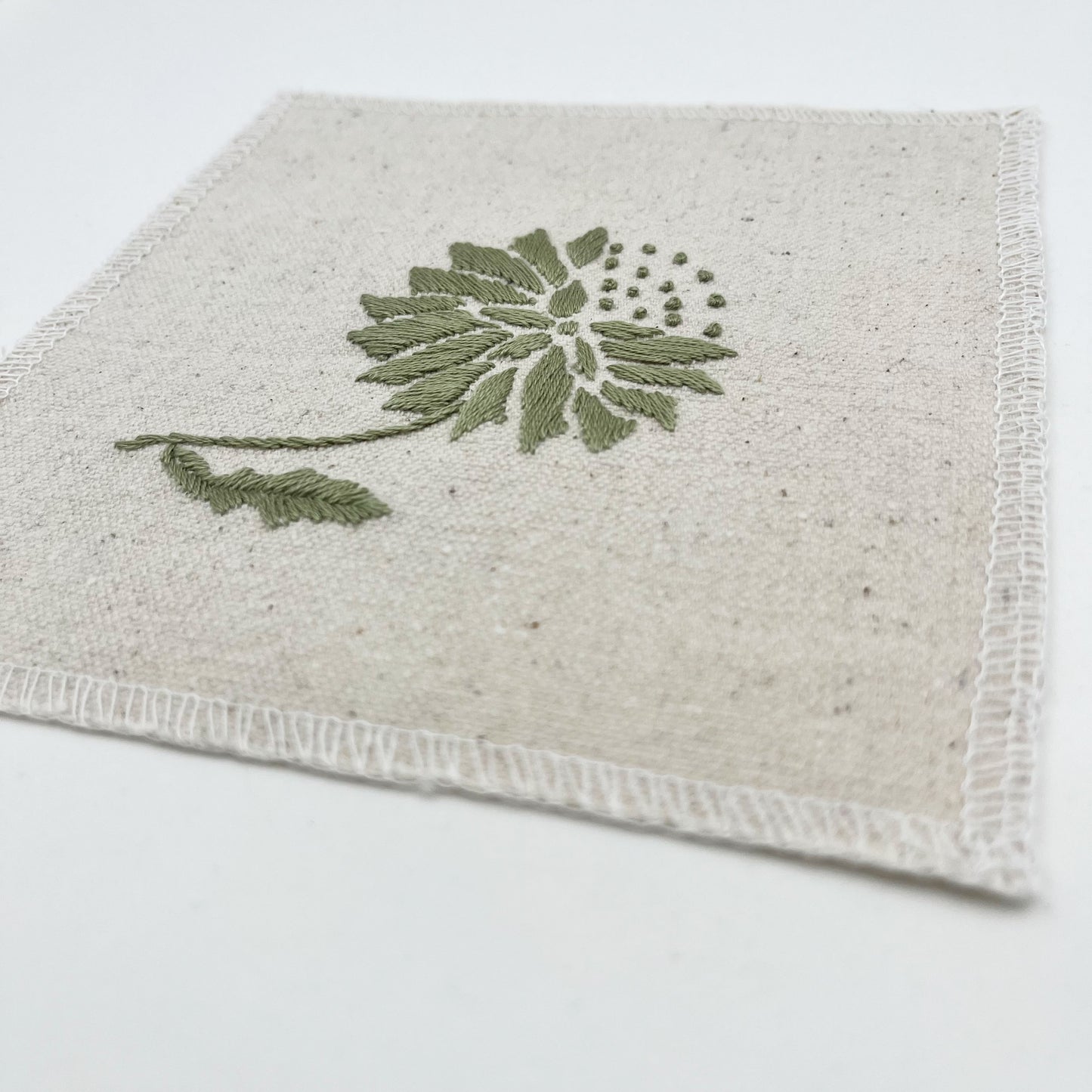a close up angled view of a square patch in natural colored fabric, hand embroidered with a sage colored abstract dandelion made mostly of petals, with a quarter of them replaced with french knots, with overlocked edges, on a white background