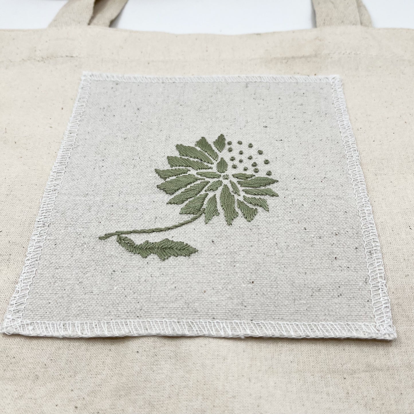 a square patch in natural colored fabric, hand embroidered with a sage colored abstract dandelion made mostly of petals, with a quarter of them replaced with french knots, with overlocked edges, on a natural colored tote bag