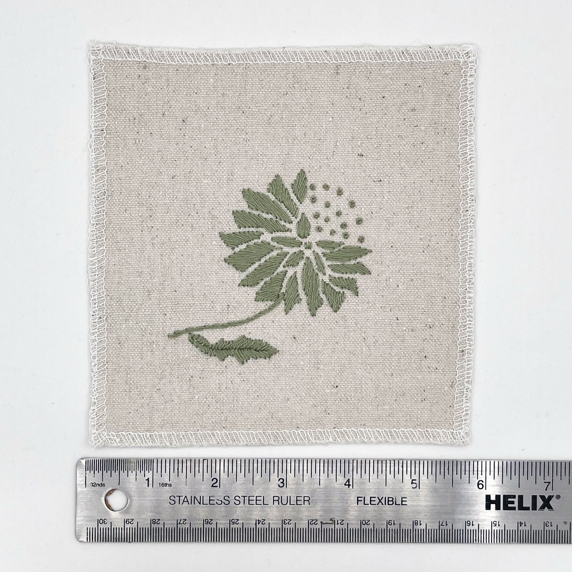 a square patch in natural colored fabric, hand embroidered with a sage colored abstract dandelion made mostly of petals, with a quarter of them replaced with french knots, with overlocked edges, next to a metal ruler to show a width of six inches