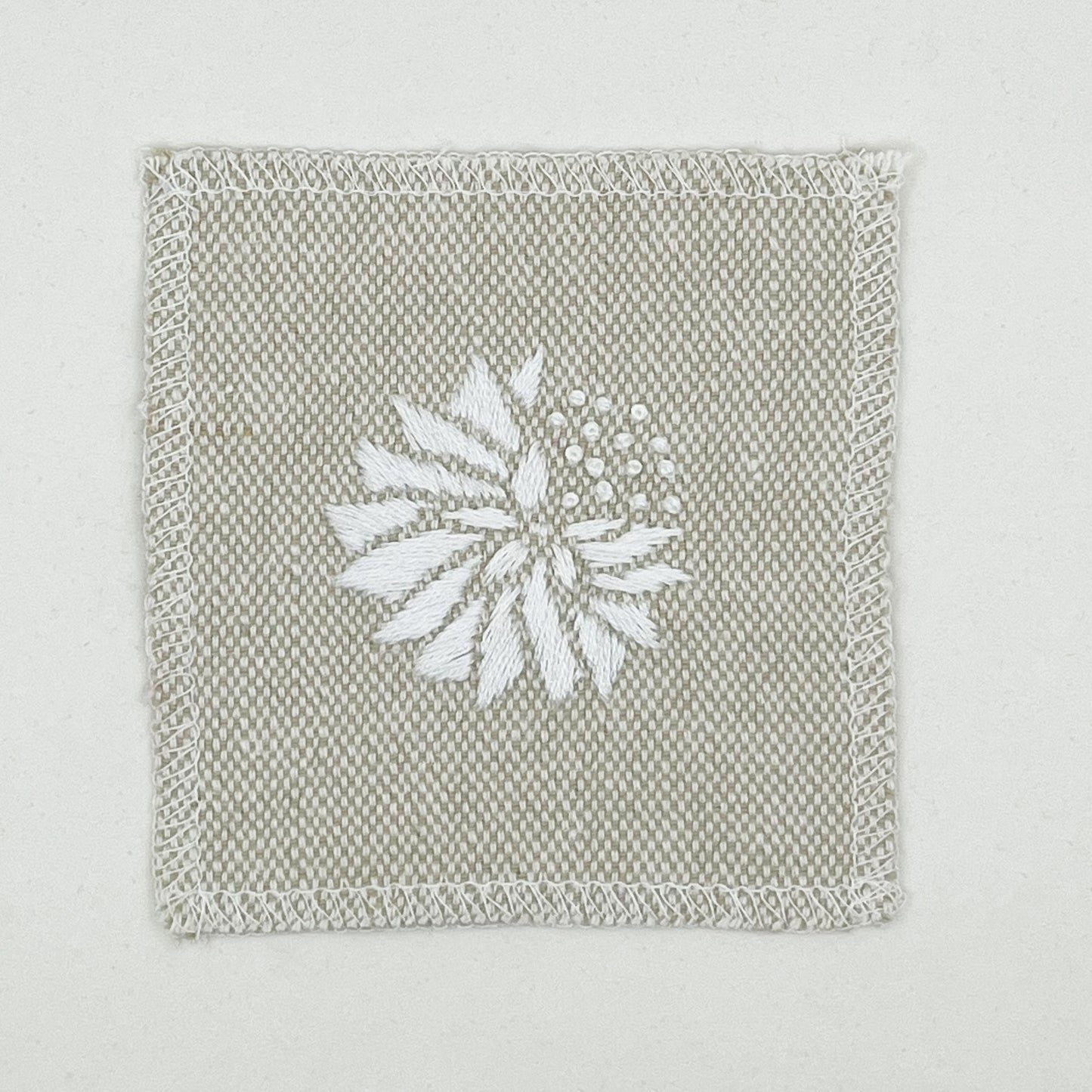 a square patch in cream fabric, hand embroidered with a white abstract dandelion made mostly of petals, with a quarter of them replaced with french knots, with overlocked edges, on a white background