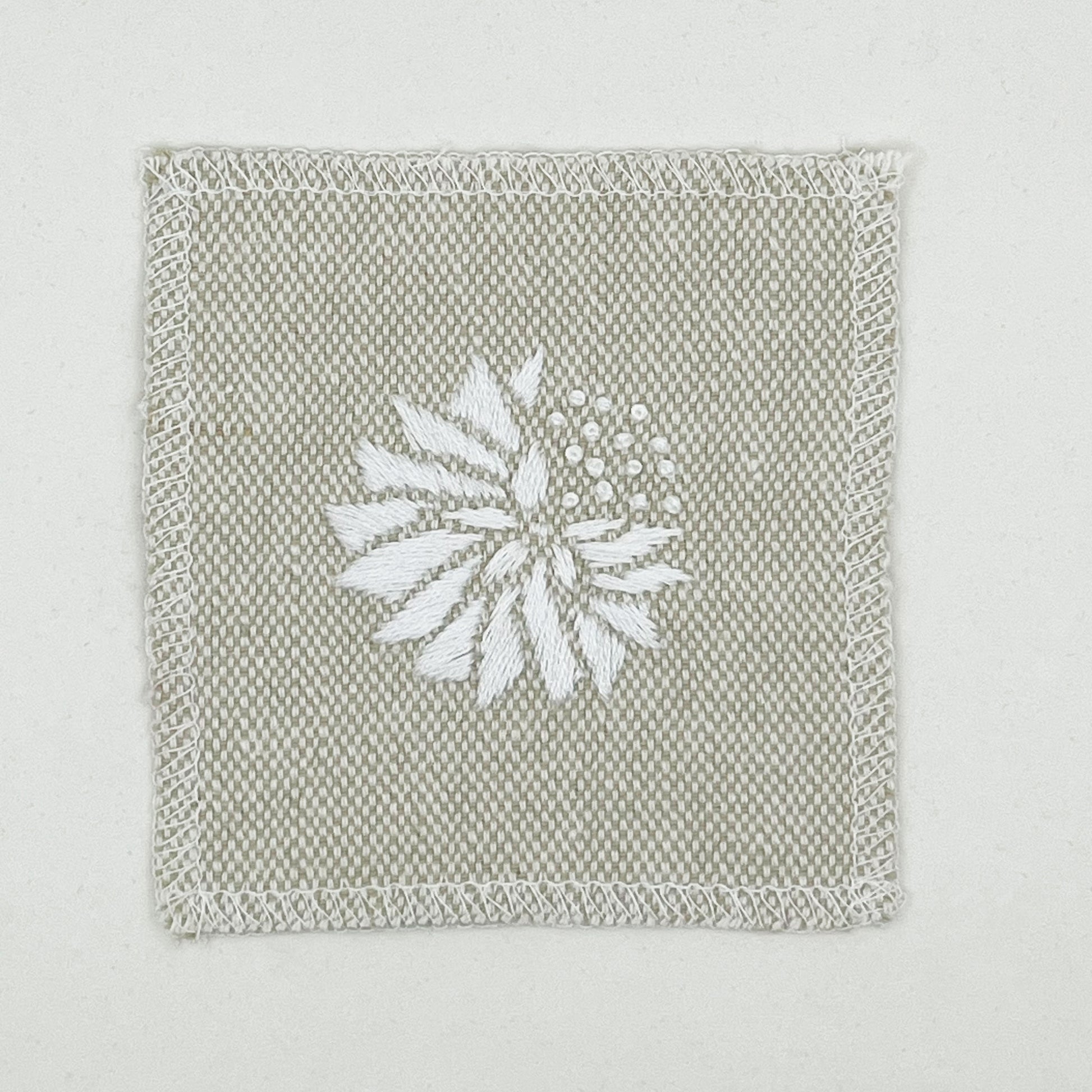 a square patch in cream fabric, hand embroidered with a white abstract dandelion made mostly of petals, with a quarter of them replaced with french knots, with overlocked edges, on a white background