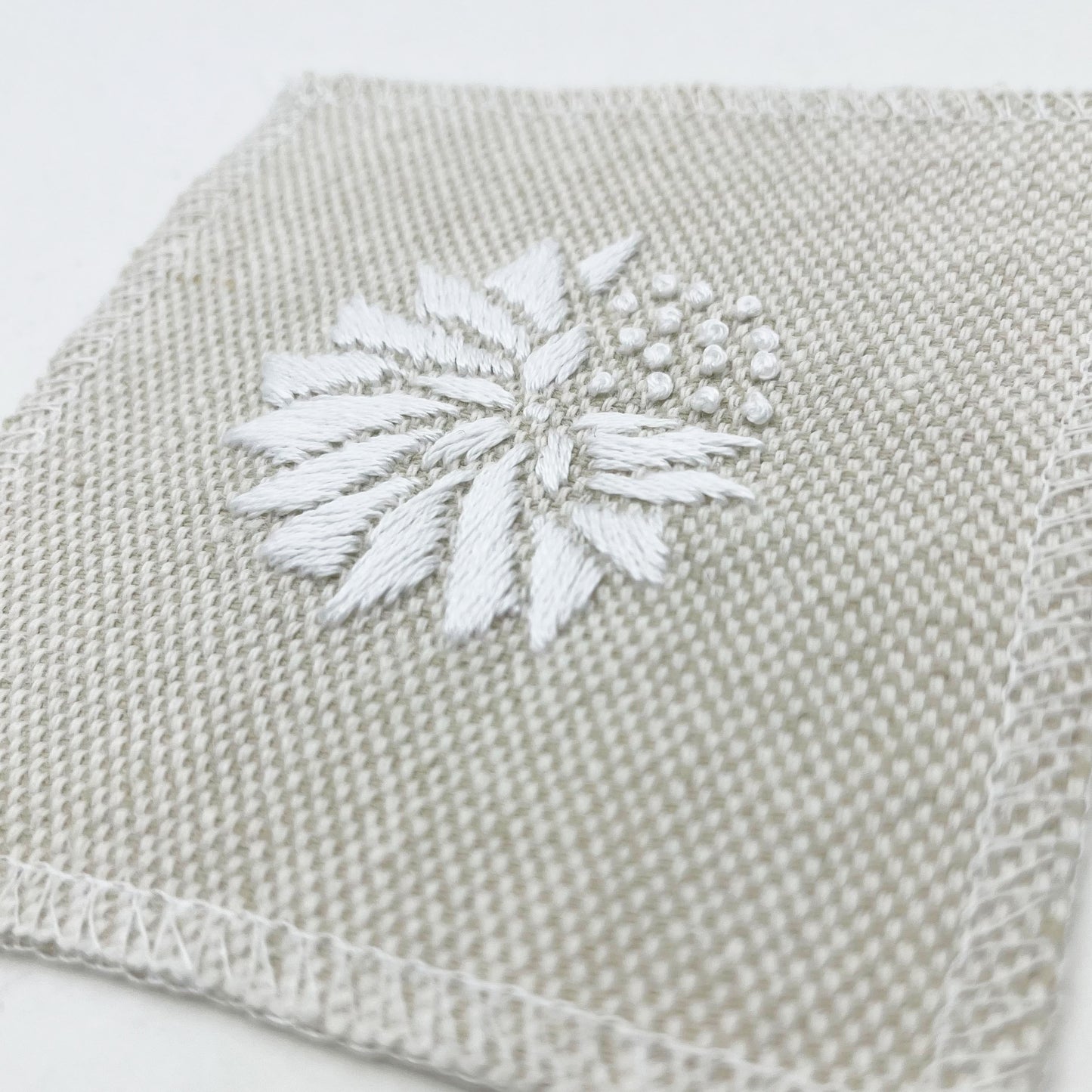 a close up angled view of a square patch in cream fabric, hand embroidered with a white abstract dandelion made mostly of petals, with a quarter of them replaced with french knots, with overlocked edges, on a white background