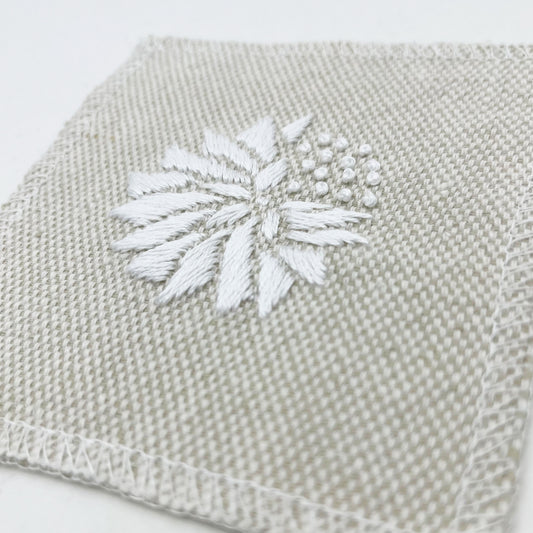 a close up angled view of a square patch in cream fabric, hand embroidered with a white abstract dandelion made mostly of petals, with a quarter of them replaced with french knots, with overlocked edges, on a white background