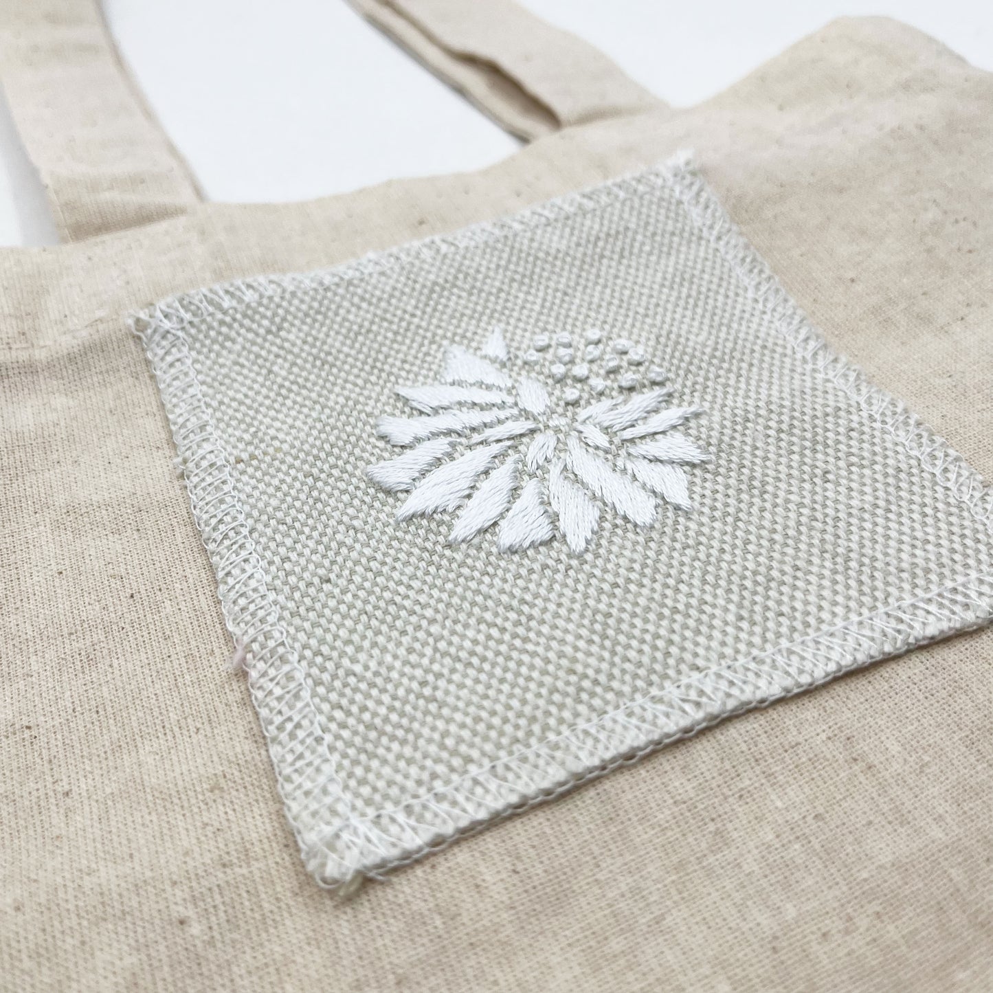 a close up angled view of a square patch in cream fabric, hand embroidered with a white abstract dandelion made mostly of petals, with a quarter of them replaced with french knots, on a matural canvas tote bag