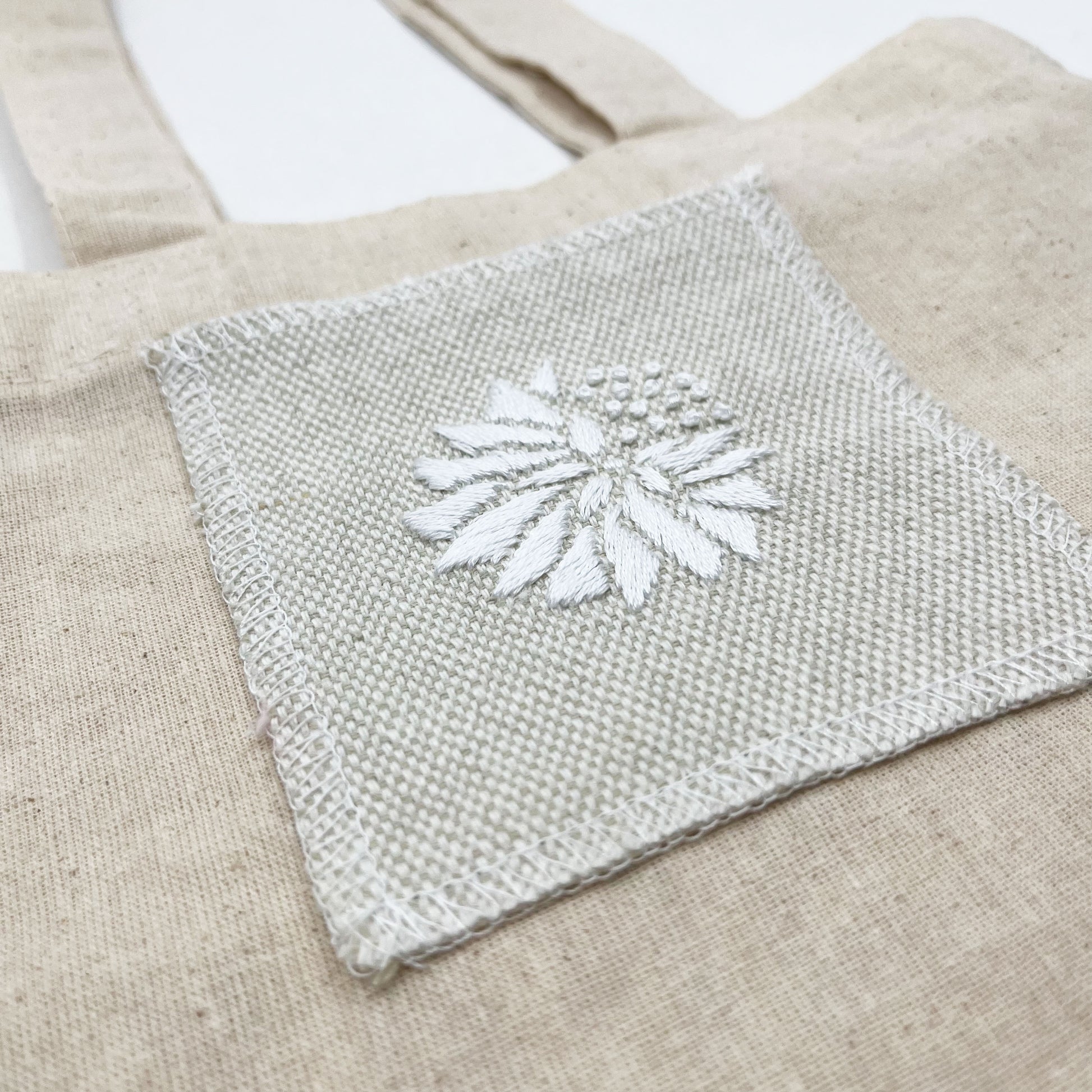 a close up angled view of a square patch in cream fabric, hand embroidered with a white abstract dandelion made mostly of petals, with a quarter of them replaced with french knots, on a matural canvas tote bag