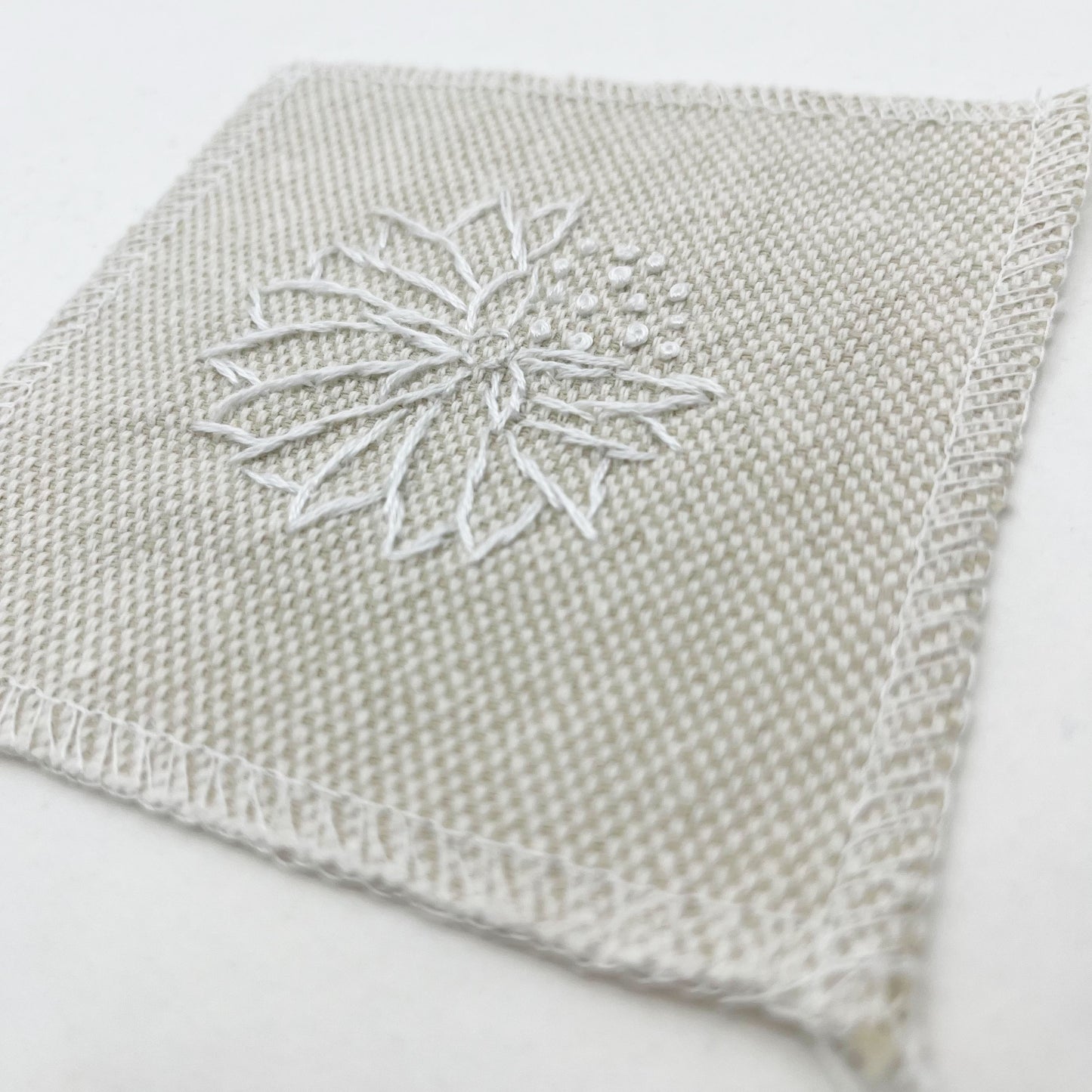 a close up angled view of a square patch in cream fabric, hand embroidered in white with the outline of an abstract dandelion made mostly of petals, with a quarter of them replaced with french knots, with overlocked edges, on a white background