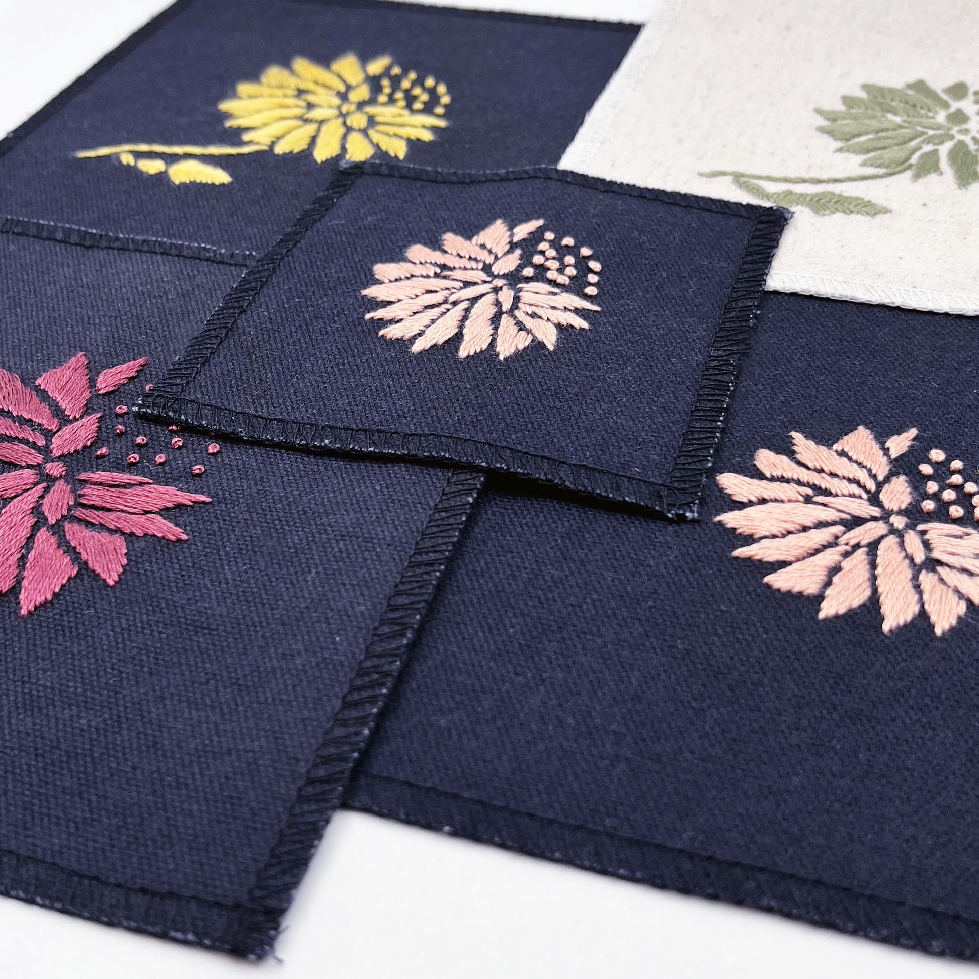 a group of square patches in natural colored and black fabric, hand embroidered with an abstract dandelion made mostly of petals, with a quarter of them replaced with french knots, in a different colors on each patch- plum, peach, sage, yellow gold
