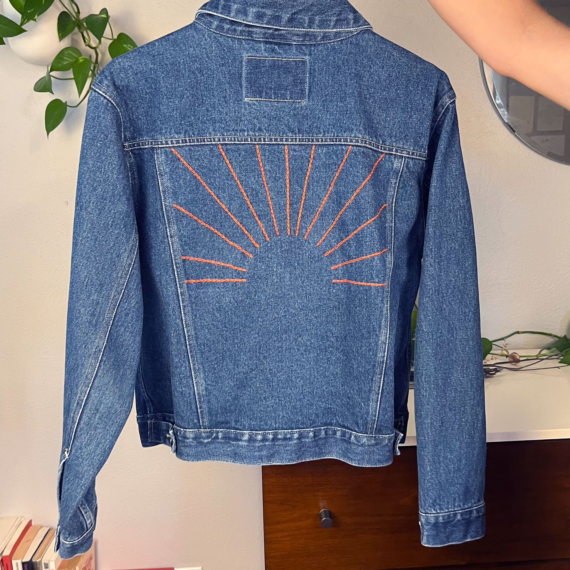  a denim jacket with hand embroidered sunbeams on the back in rust colored chainstitch, held up on a hanger