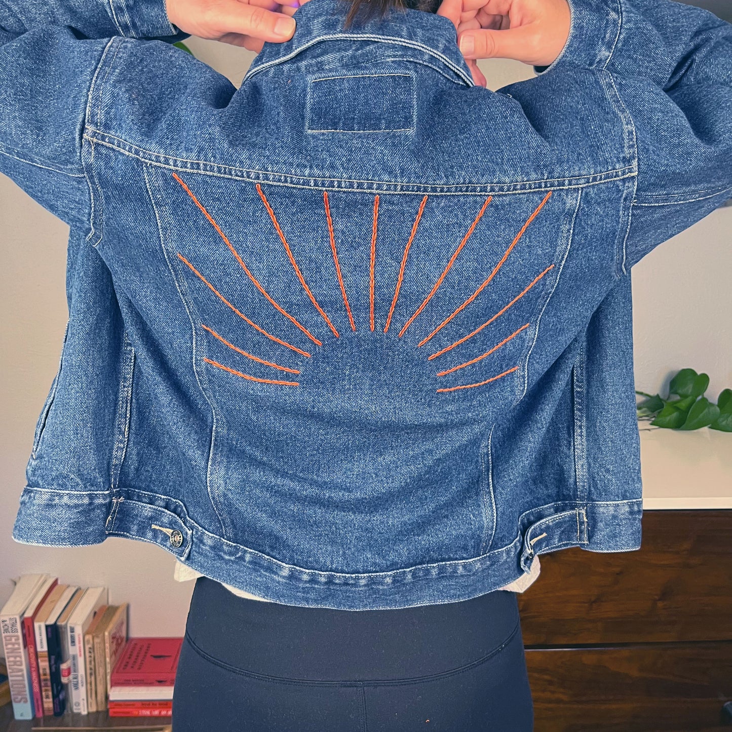 view of the back of a woman's shoulders while she adjusts the collar on a denim jacket, there are hand embroidered sunbeams on the back of the jacket in rust colored chainstitch