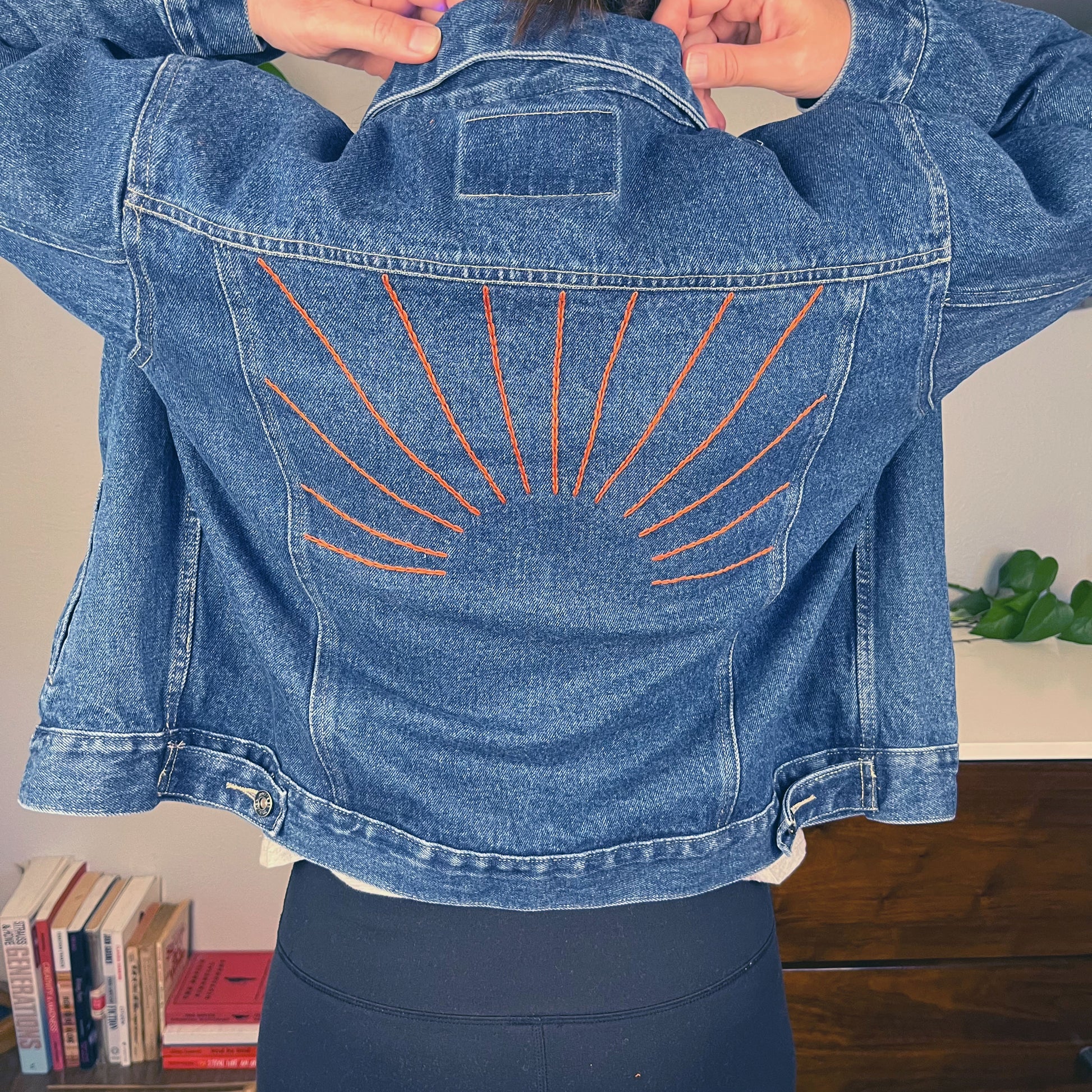 view of the back of a woman's shoulders while she adjusts the collar on a denim jacket, there are hand embroidered sunbeams on the back of the jacket in rust colored chainstitch