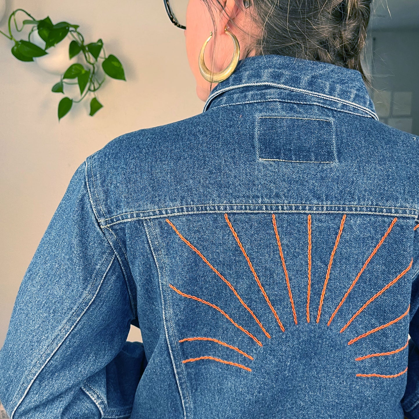 close up view of the back of a woman's shoulders, she's wearing a denim jacket with hand embroidered sunbeams on the back in rust colored chainstitch