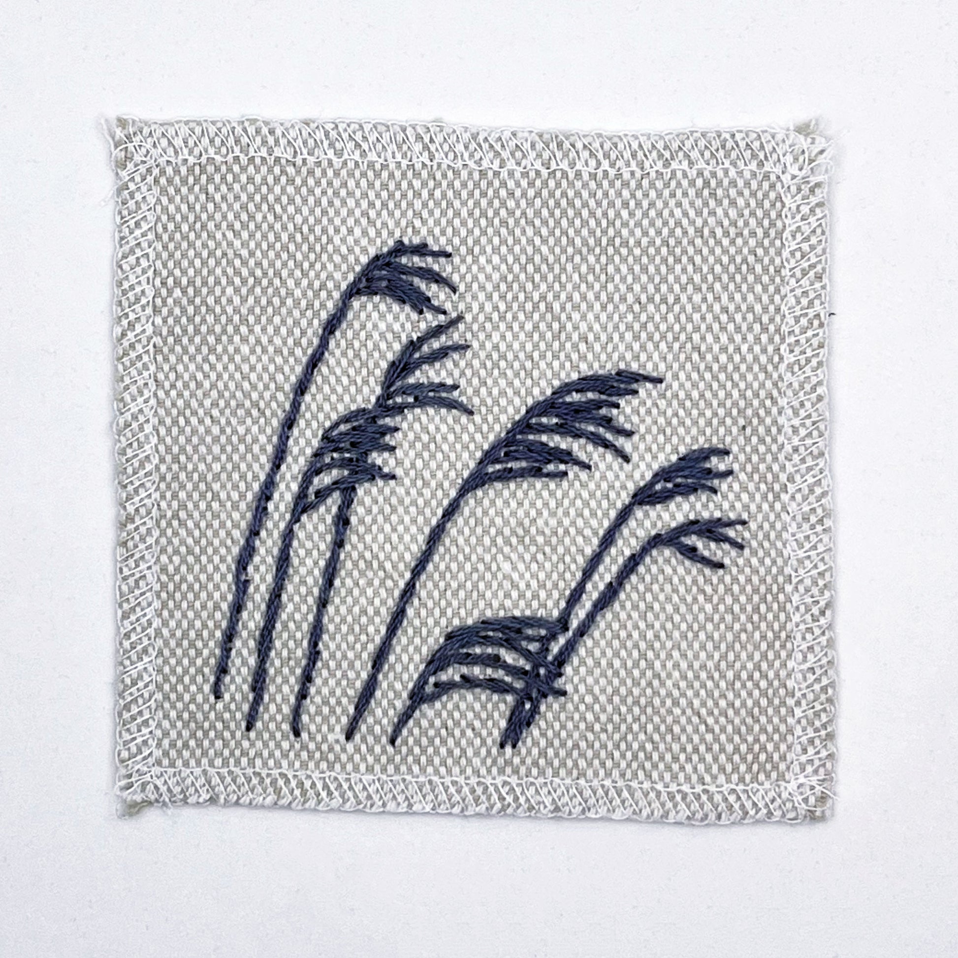 a natural colored square patch with stalks of wild grass stitched in grey thread with a stem stitch, on a white background