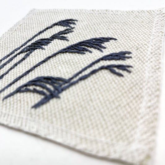 close up angled view of a natural colored square patch with stalks of wild grass stitched in grey thread with a stem stitch, on a white background