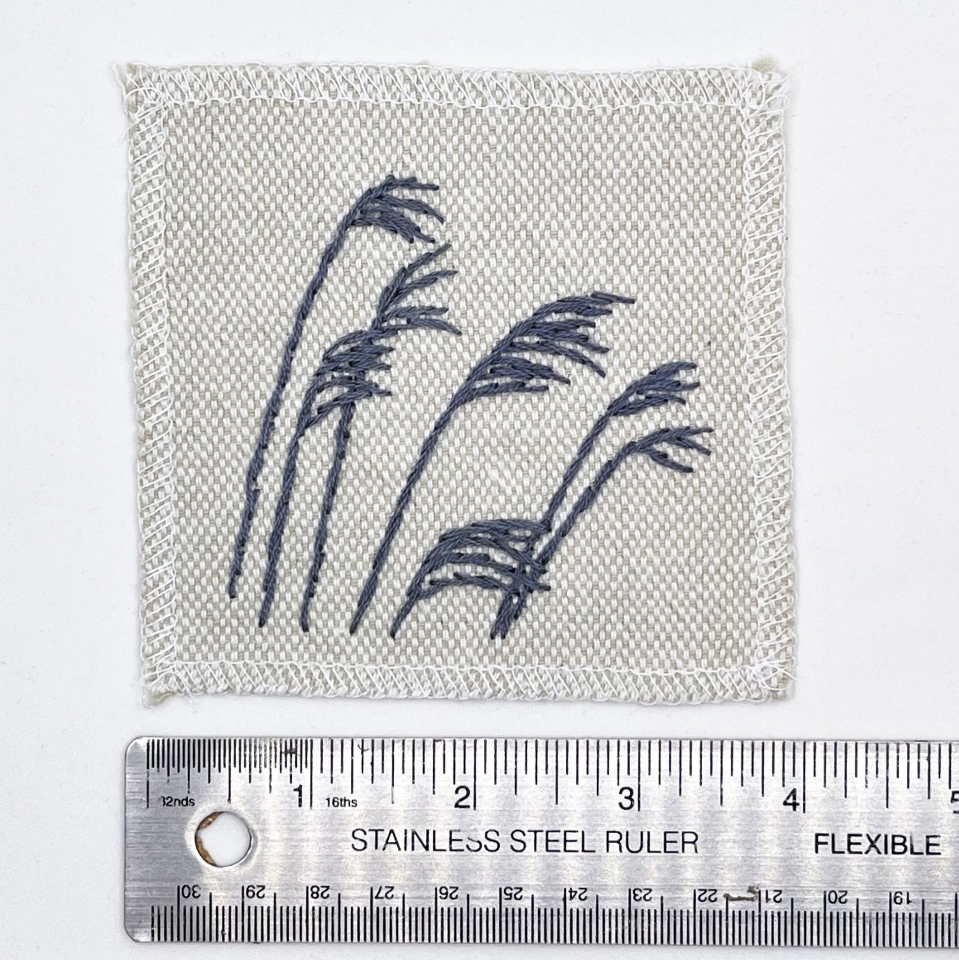 a natural colored square patch with stalks of wild grass stitched in grey thread with a stem stitch, placed next to a metal ruler to show a width of 4 inches, on a white background