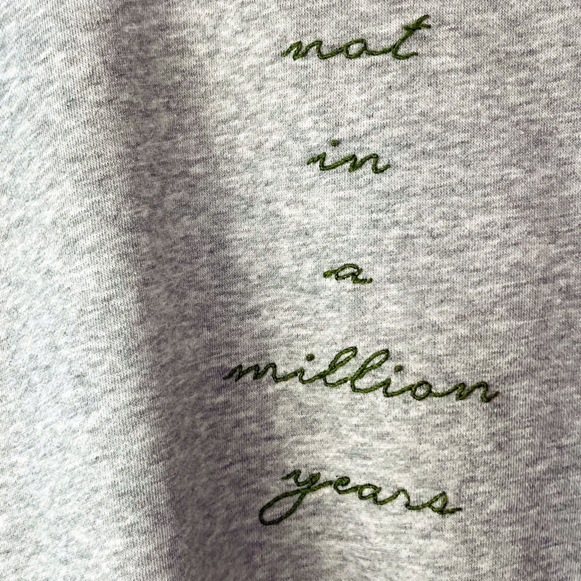 close up view of a grey raglan sweatshirt with the words not in a million years hand embroidered in pine green in cursive