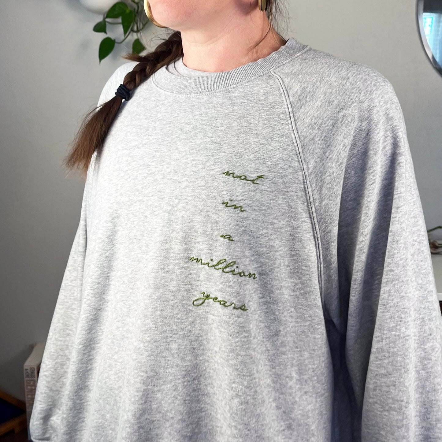 a woman wearing a grey raglan sweatshirt with the words not in a million years hand embroidered in pine green down the wearers left side