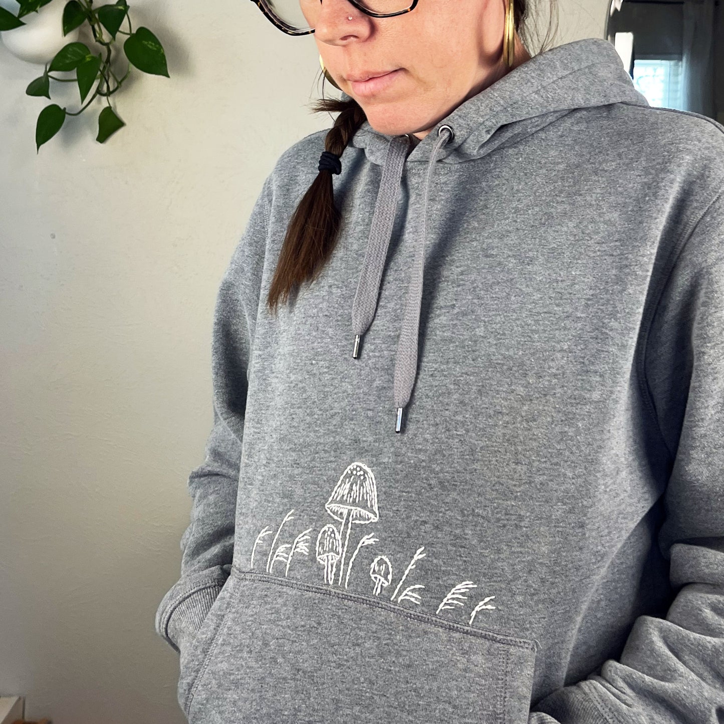 a woman wearing a grey hoodie with hand embroidered mushrooms above the pocket