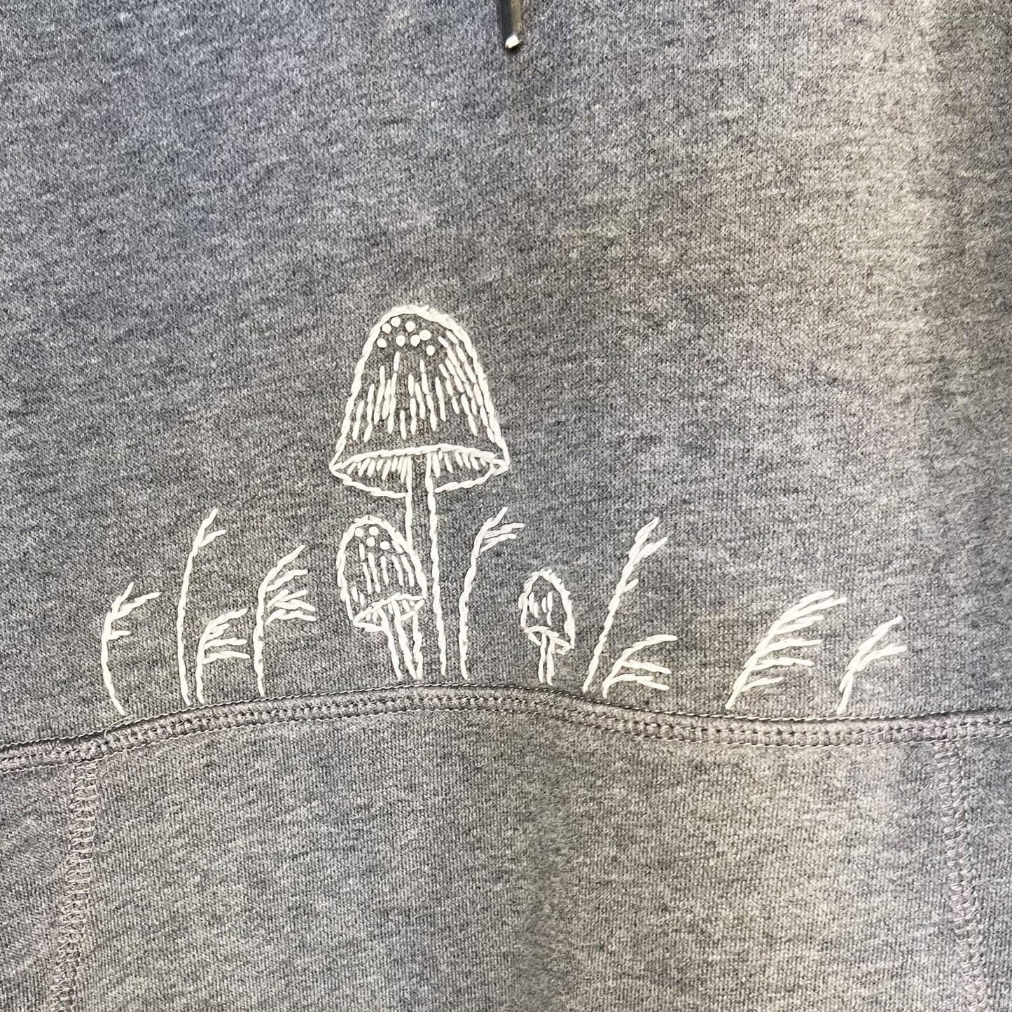 close up view of white hand embroidered mushrooms above the pocket of a grey hoodie