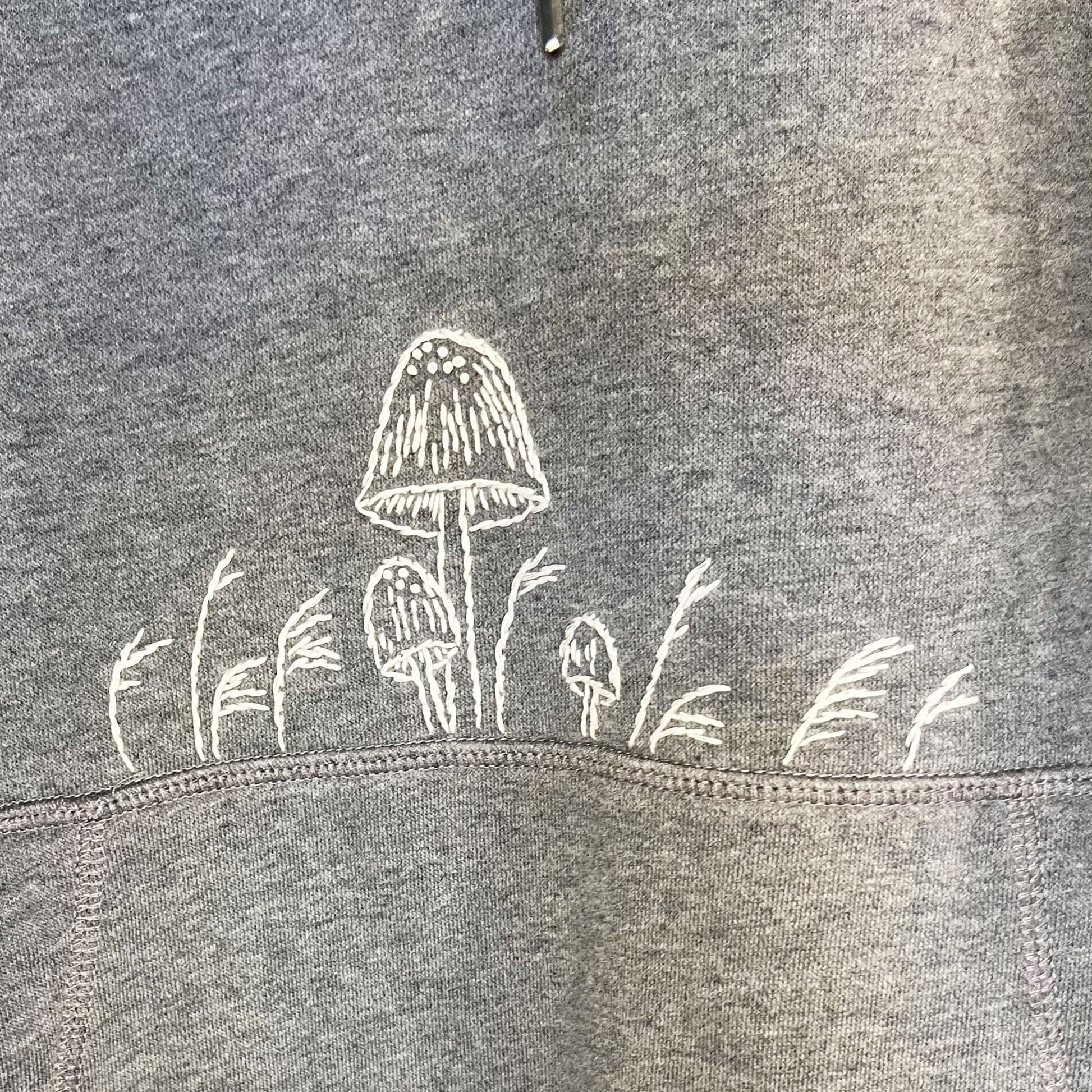 close up view of white hand embroidered mushrooms above the pocket of a grey hoodie