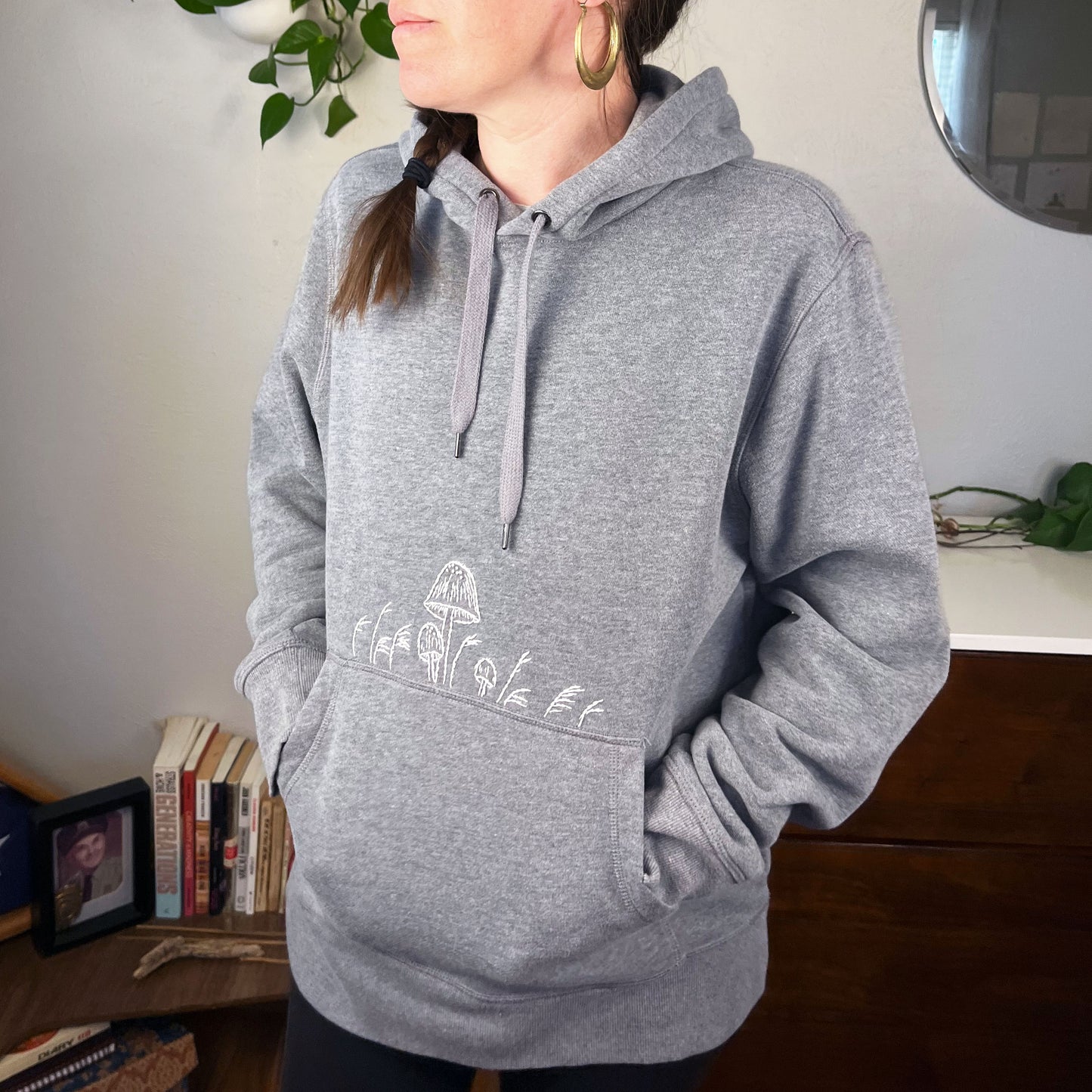 a woman wearing a grey hoodie with hand embroidered mushrooms above the pocket