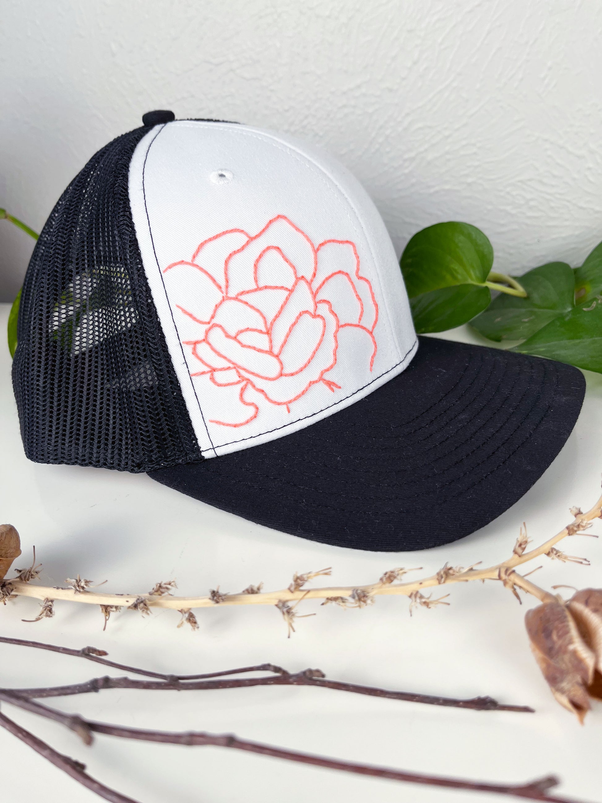 black mesh hat with neon pink outline of a peony hand embroidered to the front, sitting on a white dresser with live and dried plants around it