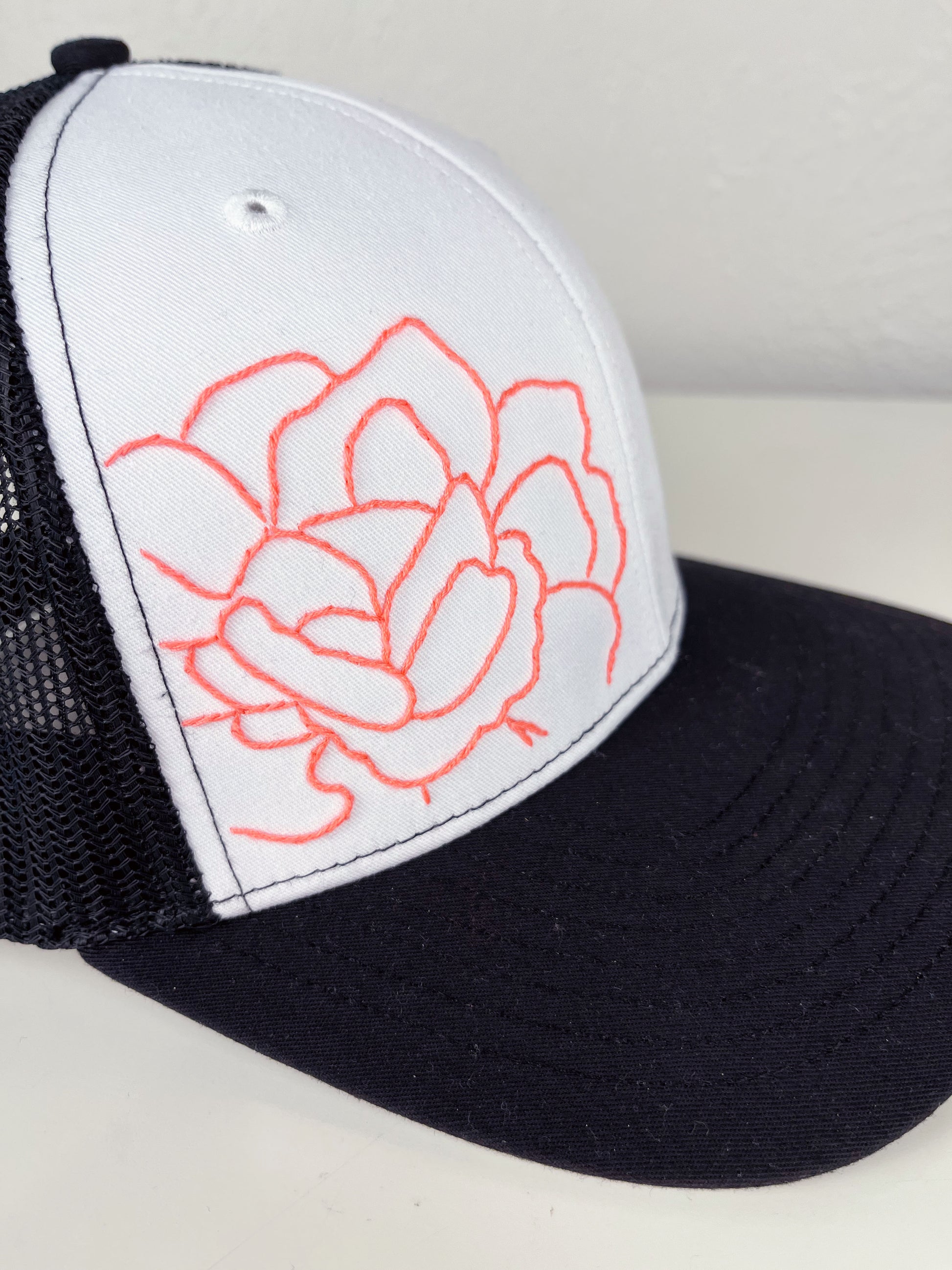close up view of a black mesh hat with neon pink outline of a peony hand embroidered to the front, sitting on a white dresser