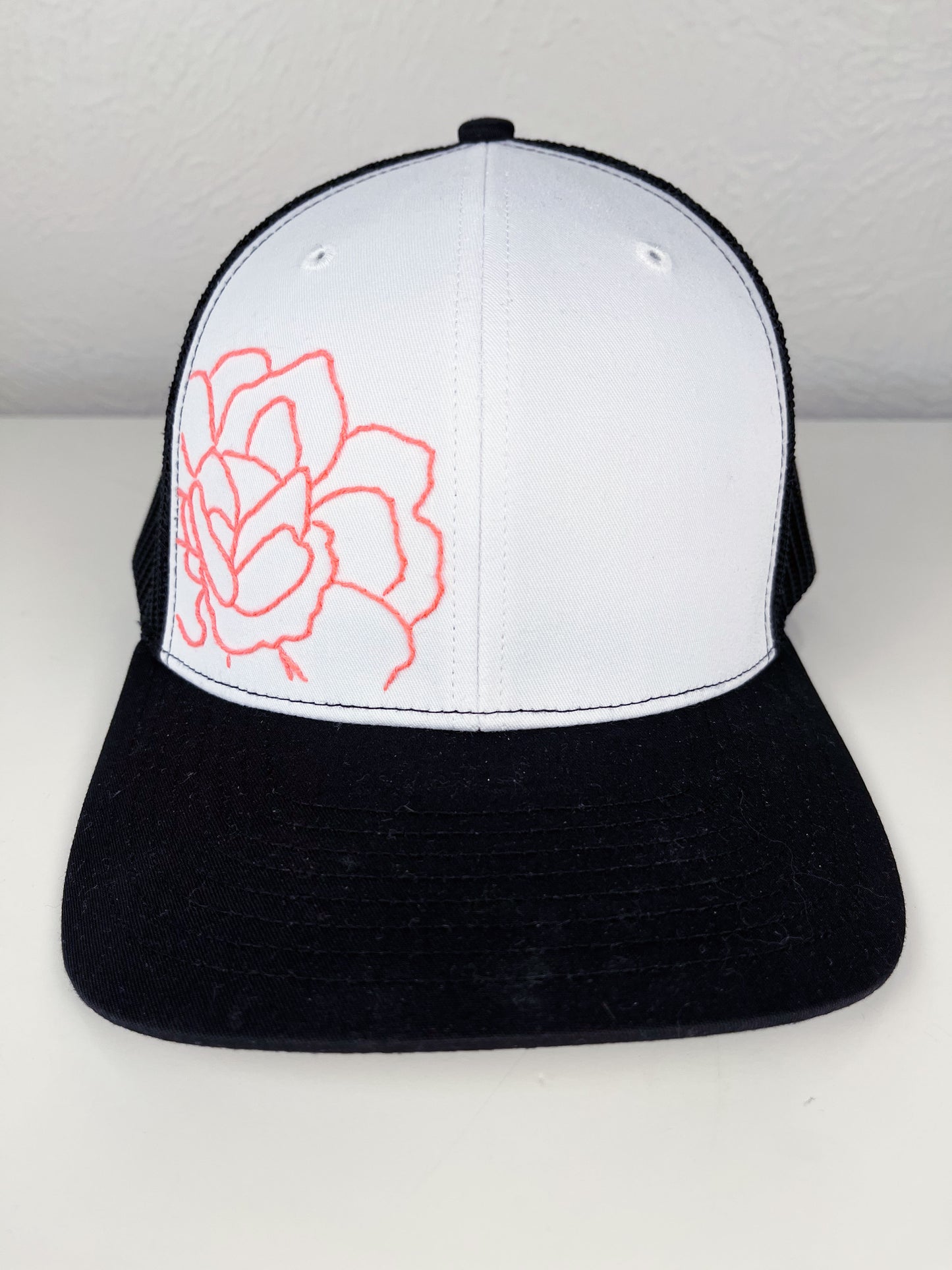 black mesh hat with neon pink outline of a peony hand embroidered to the front, sitting on a white dresser