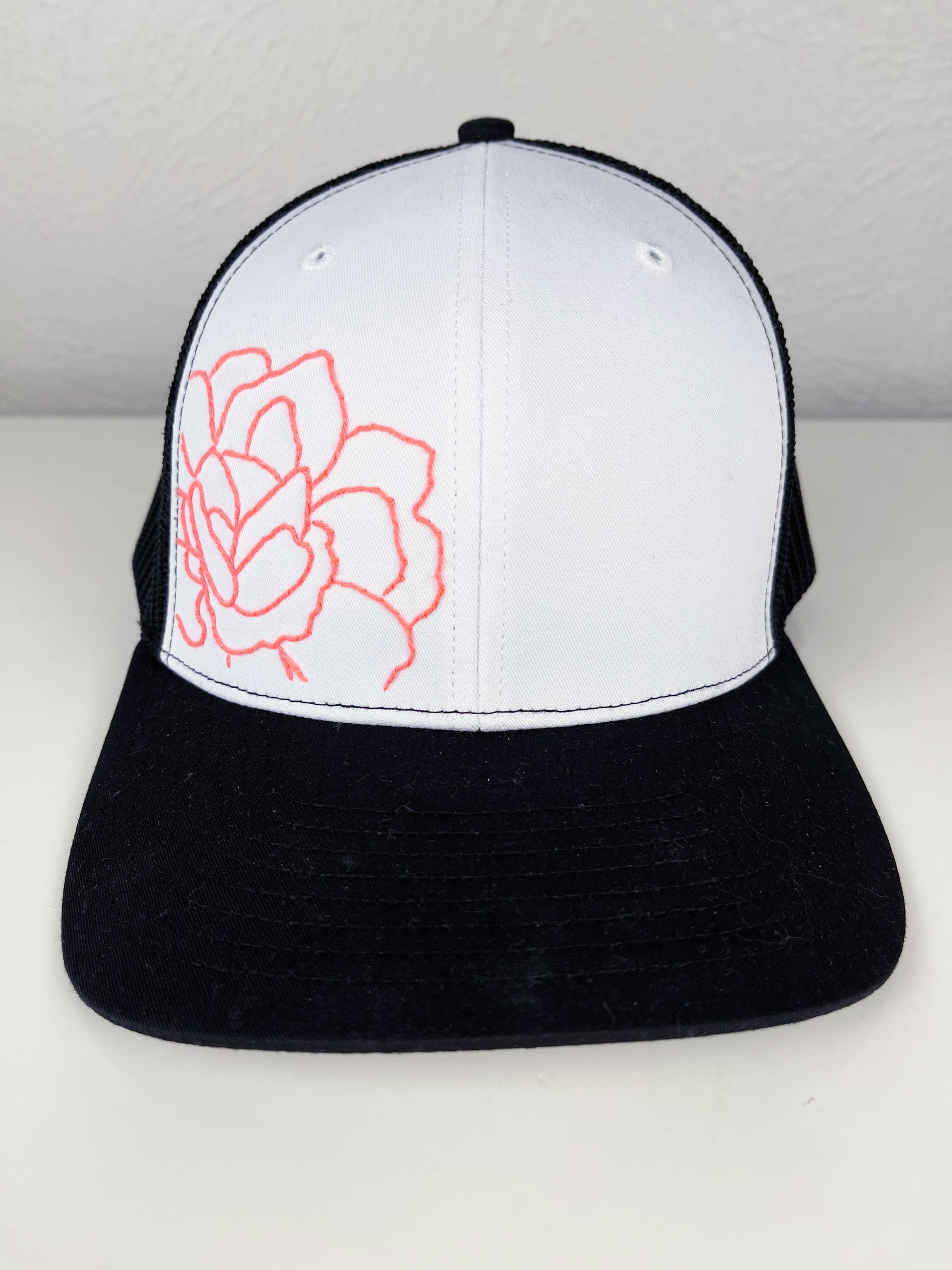 black mesh hat with neon pink outline of a peony hand embroidered to the front, sitting on a white dresser