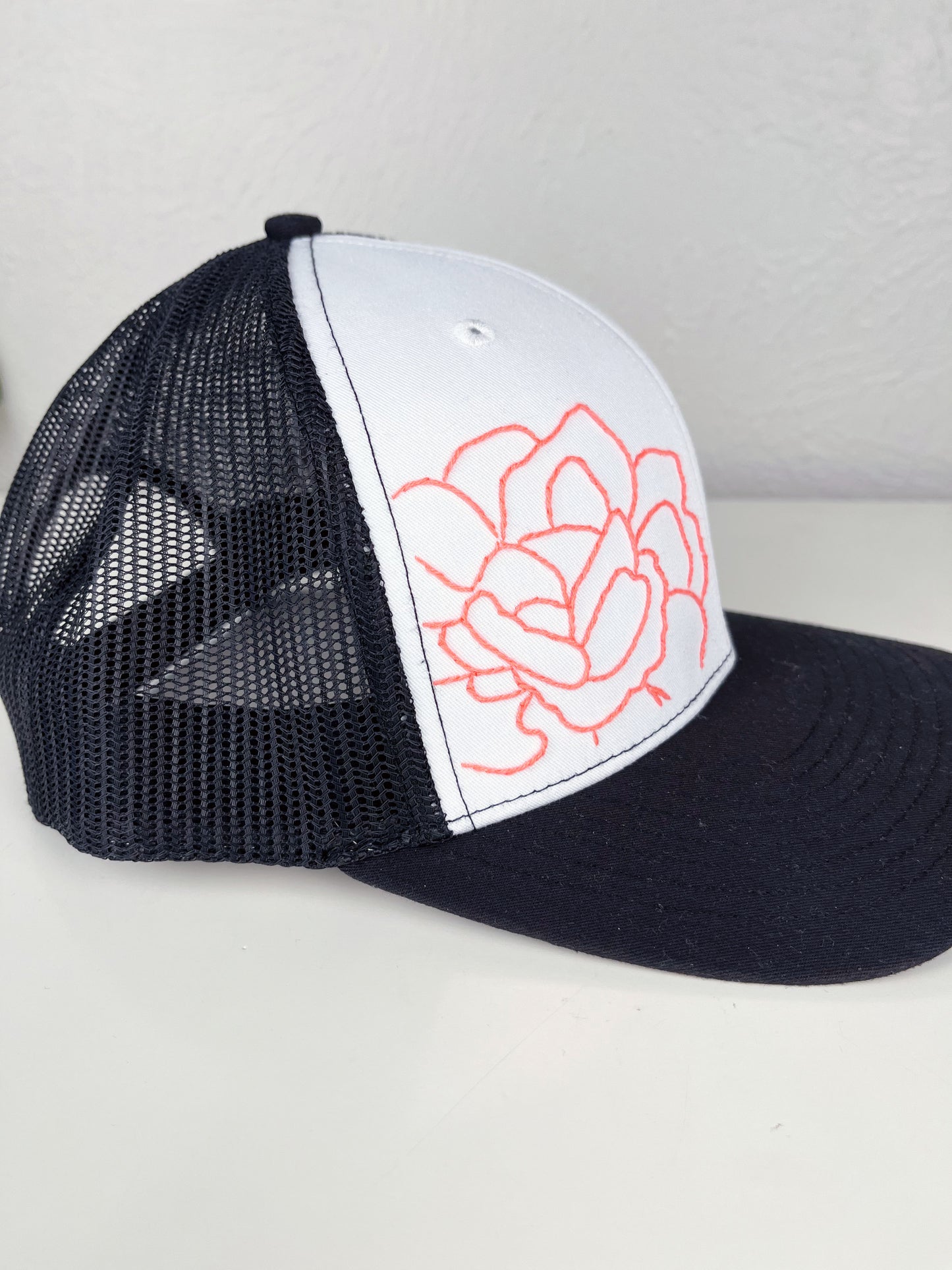 side view of black mesh hat with neon pink outline of a peony hand embroidered to the front, sitting on a white dresser