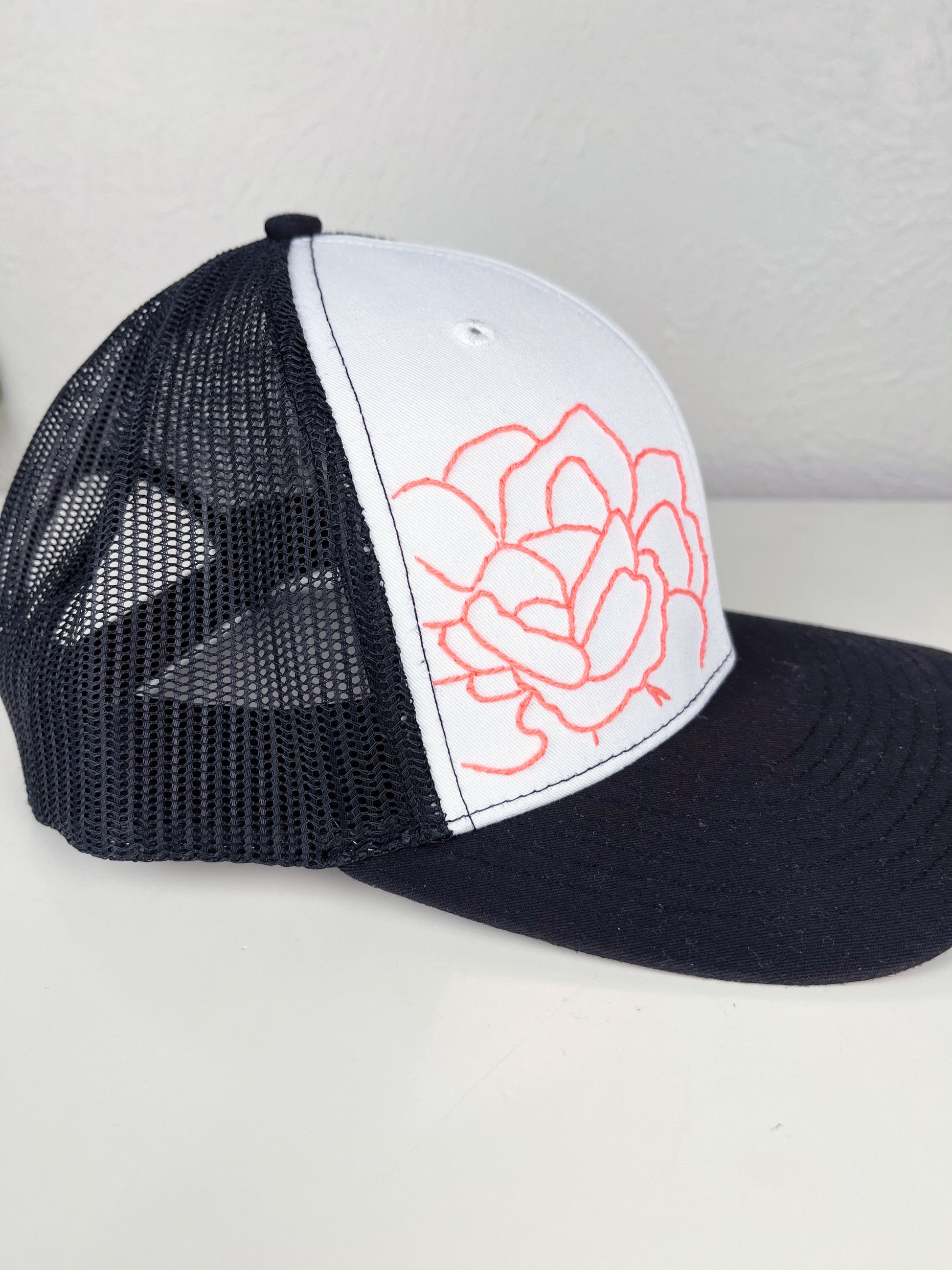 side view of black mesh hat with neon pink outline of a peony hand embroidered to the front, sitting on a white dresser