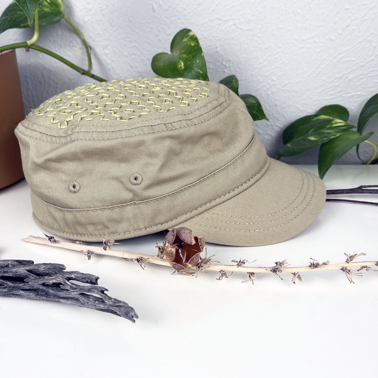 tan cadet hat with persimmon sashiko pattern stitched in chartreuse thread on the top, sitting on a dresser with a plant vine and dried flowers around it