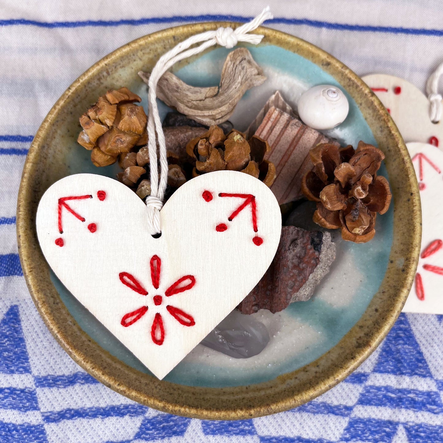 laser cut wood heart ornament embellished with hand embroidery in red sitting on a dish of found nature objects on a cream and blue patterned scarf