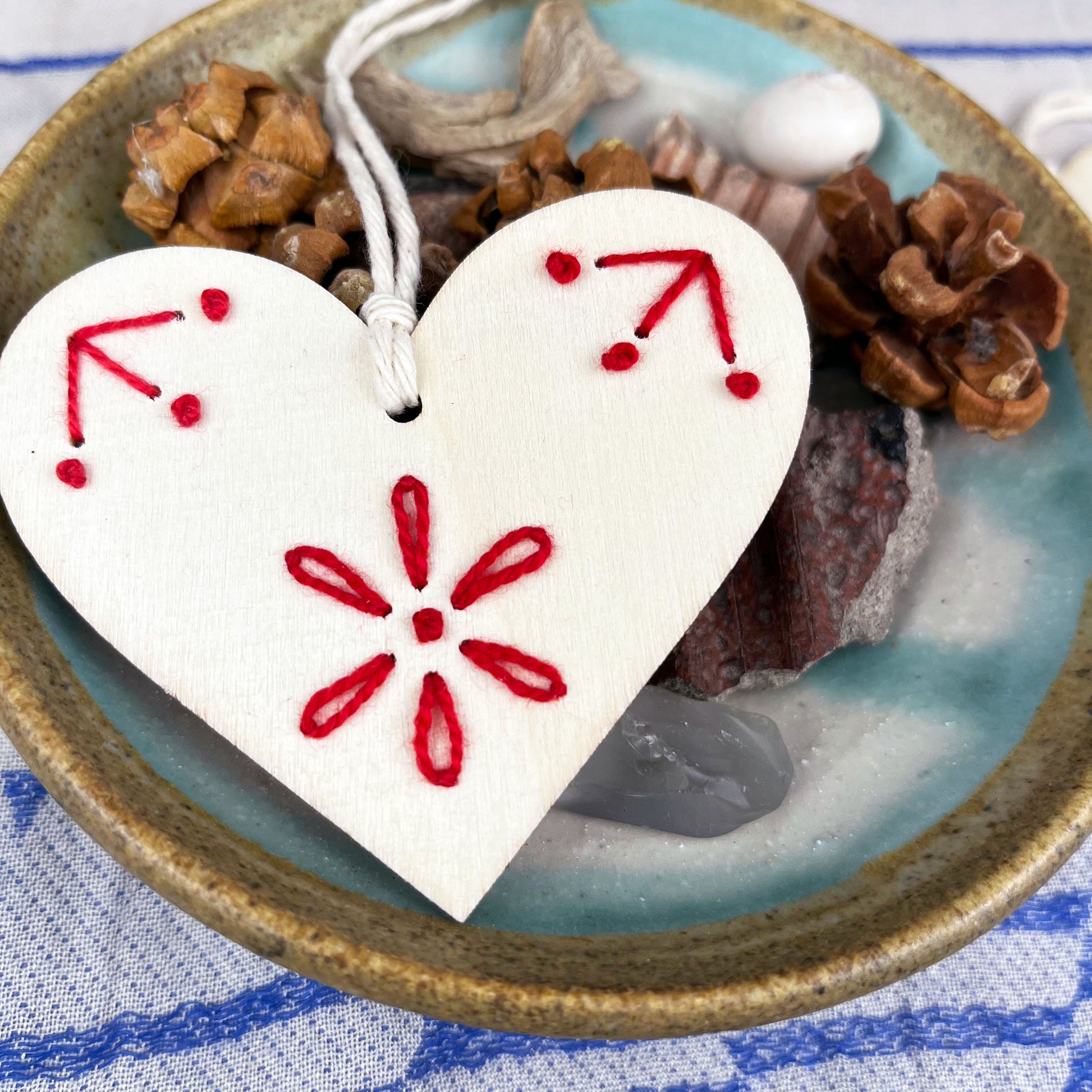laser cut wood heart ornament embellished with hand embroidery in red sitting on a dish of found nature objects on a cream and blue patterned scarf