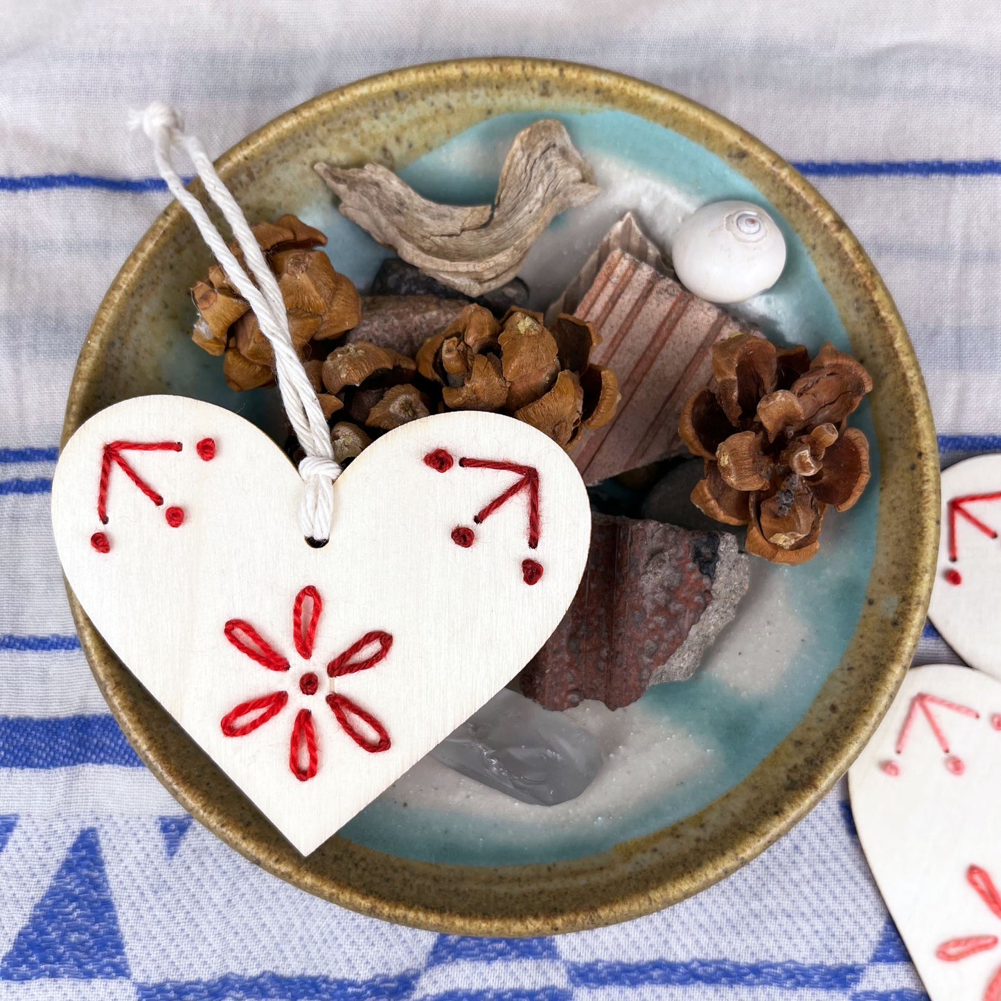 laser cut wood heart ornament embellished with hand embroidery in red sitting on a dish of found nature objects on a cream and blue patterned scarf