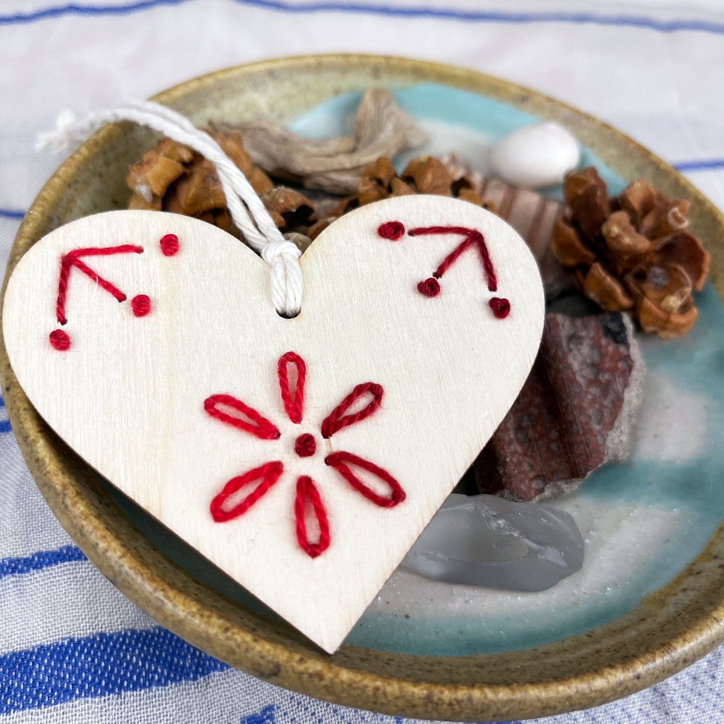 laser cut wood heart ornament embellished with hand embroidery in red sitting on a dish of found nature objects on a cream and blue patterned scarf