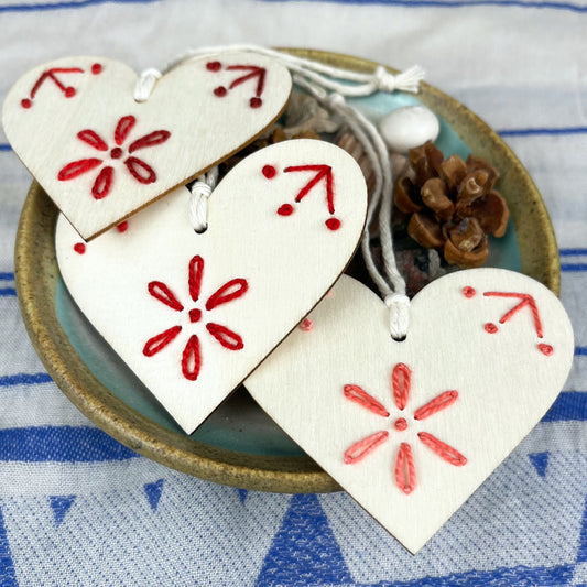 three laser cut wood heart ornaments embellished with hand embroidery in pink and reds sitting on a dish of found nature objects on a cream and blue patterned scarf