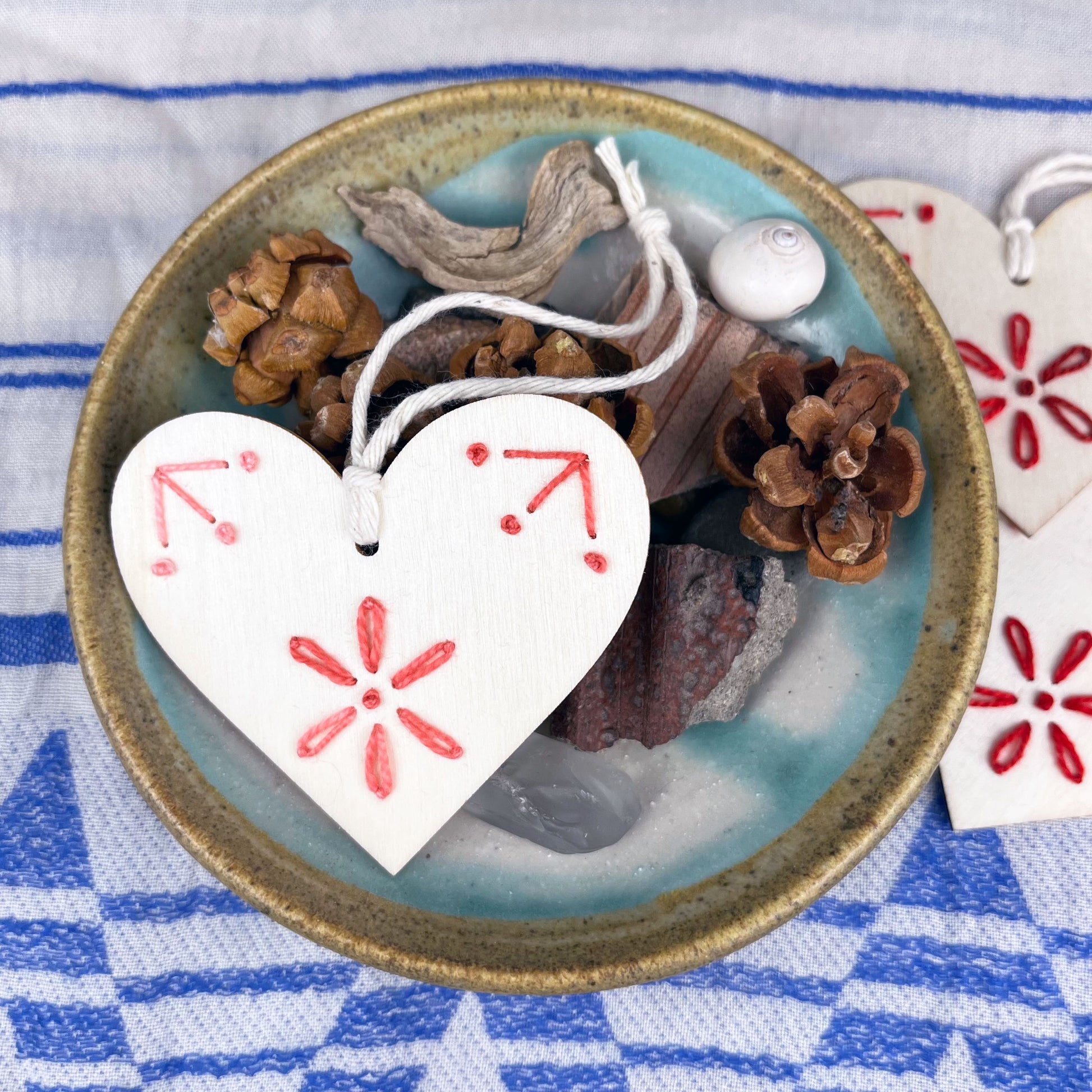 laser cut wood heart ornament embellished with hand embroidery in pink sitting on a dish of found nature objects on a cream and blue patterned scarf